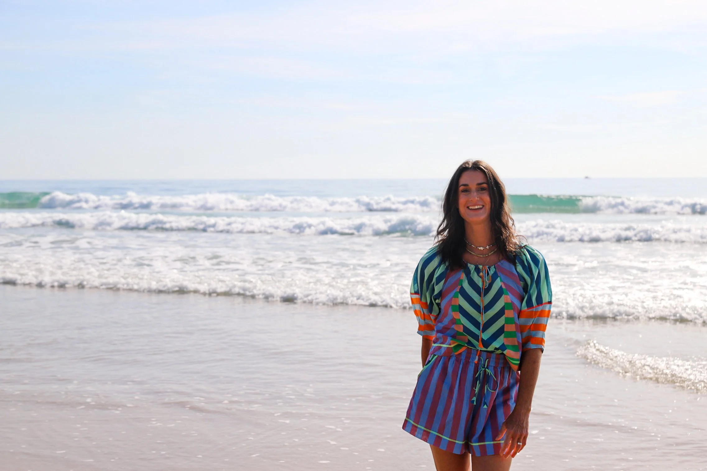 A woman with dark brown hair wearing a colorful striped dress standing on the beach with waves in the background.
