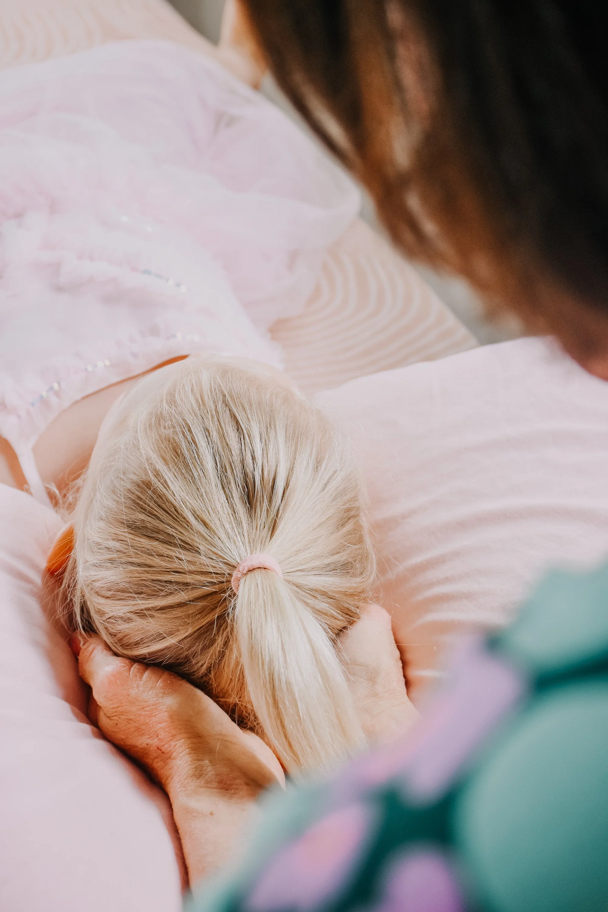 Close-up of a person with light blonde hair holding a tiny, sleeping baby with platinum blonde hair. The person is cradling the baby's head with their hand.