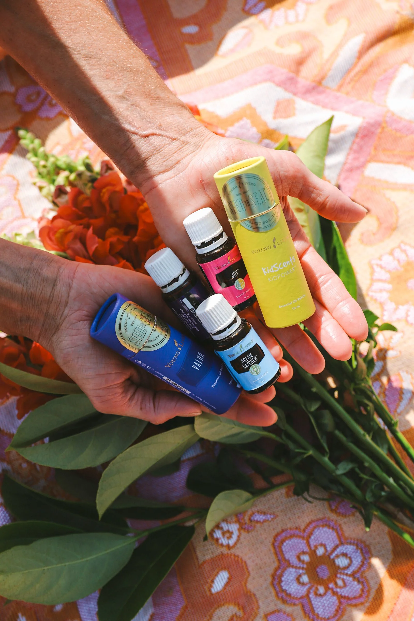 Several essential oil bottles are held in hands above a flower and leaf arrangement on a colorful tablecloth.