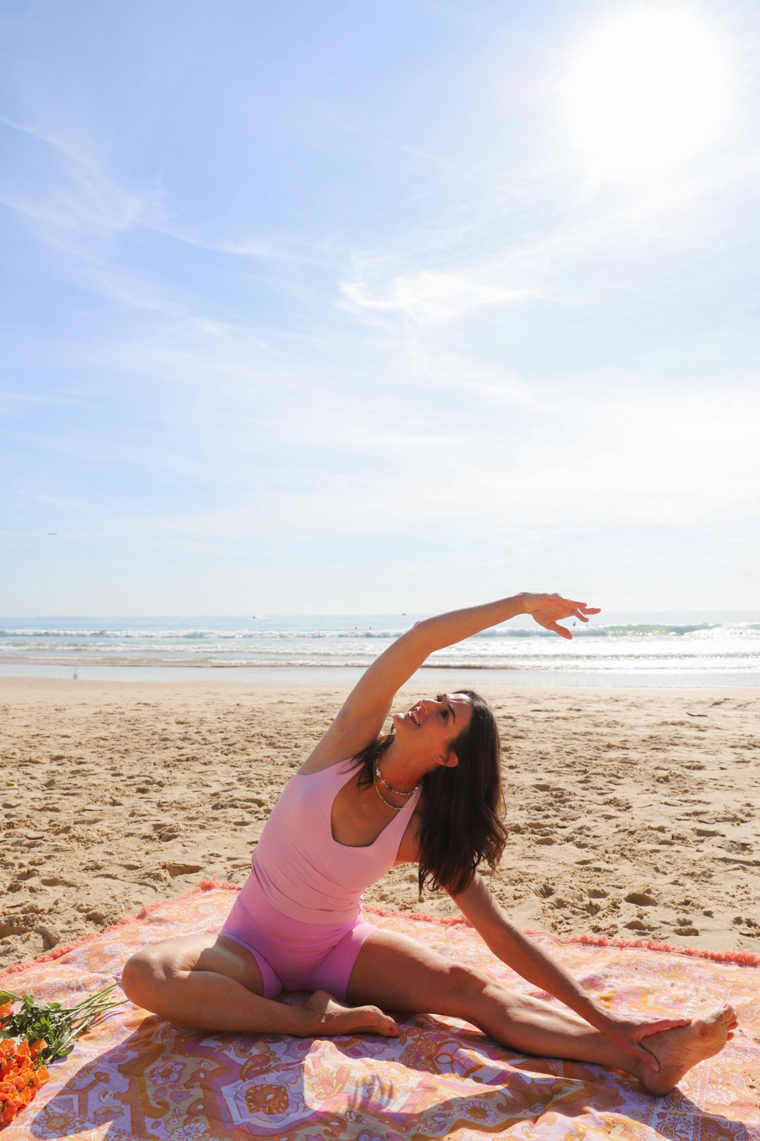 A woman practicing yoga on a beach during daytime, sitting on a pink and orange blanket, with the ocean and sky in the background, stretching her arm overhead.