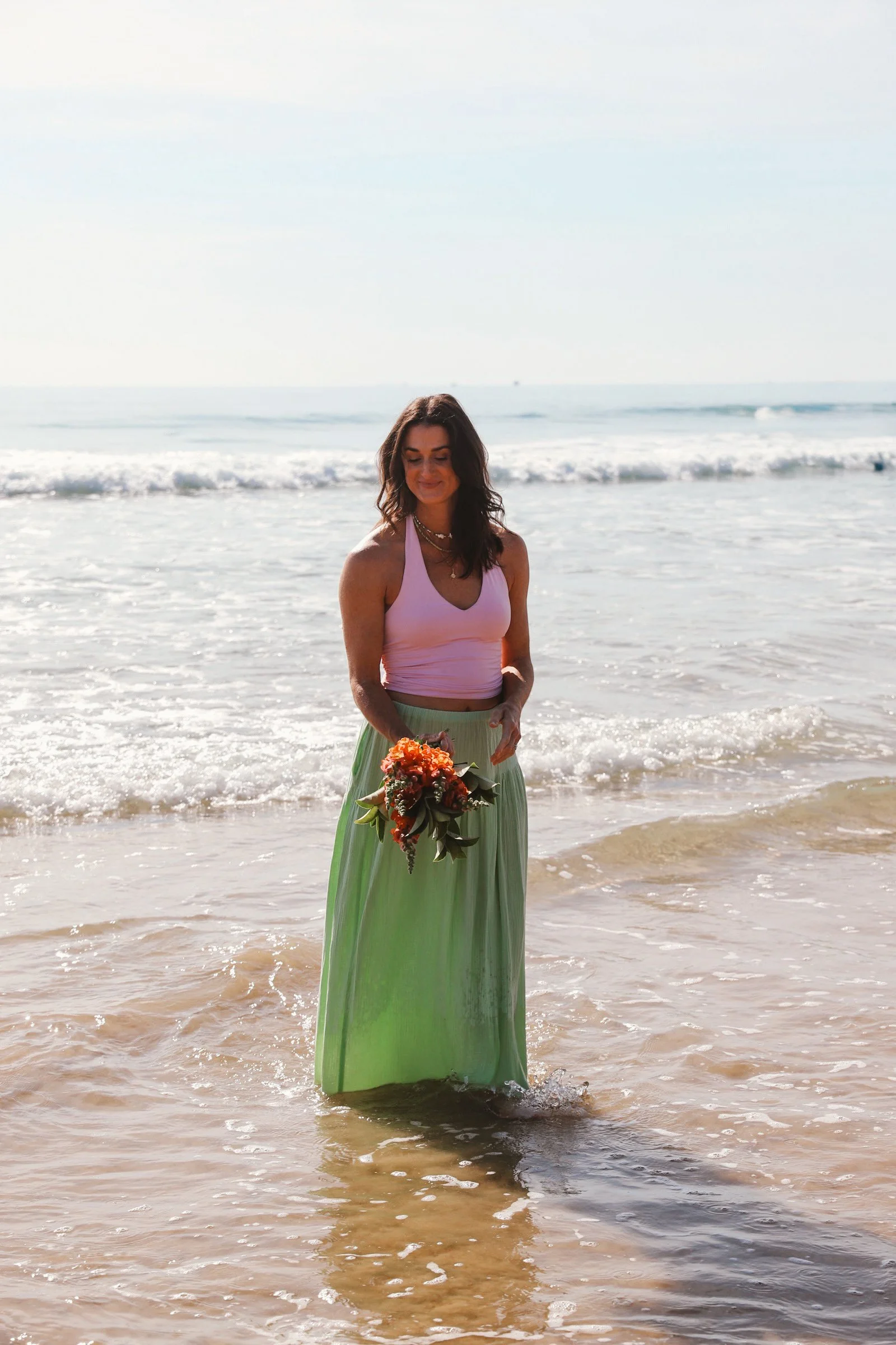 A woman in a flowing green skirt and pink top standing in shallow ocean water, holding a bouquet of orange and red flowers, with waves and clear sky in the background.
