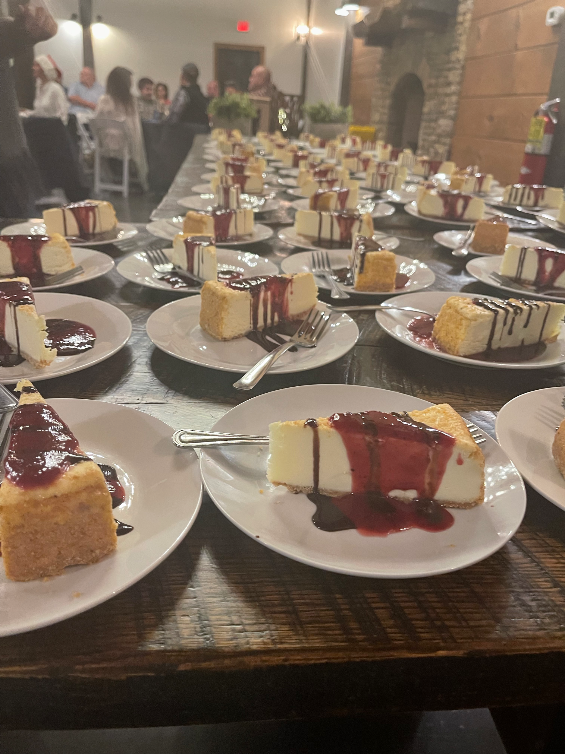 Multiple plates of cheesecake with berry sauce on a long wooden table in a restaurant setting.