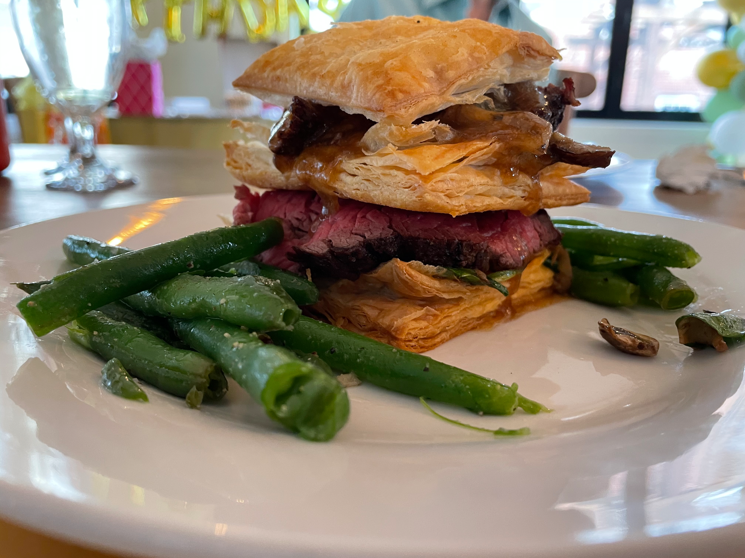 Close-up of a dish with cooked green beans, rare sliced beef, and a flaky pastry in a white dish, with a background of a decorated restaurant interior.