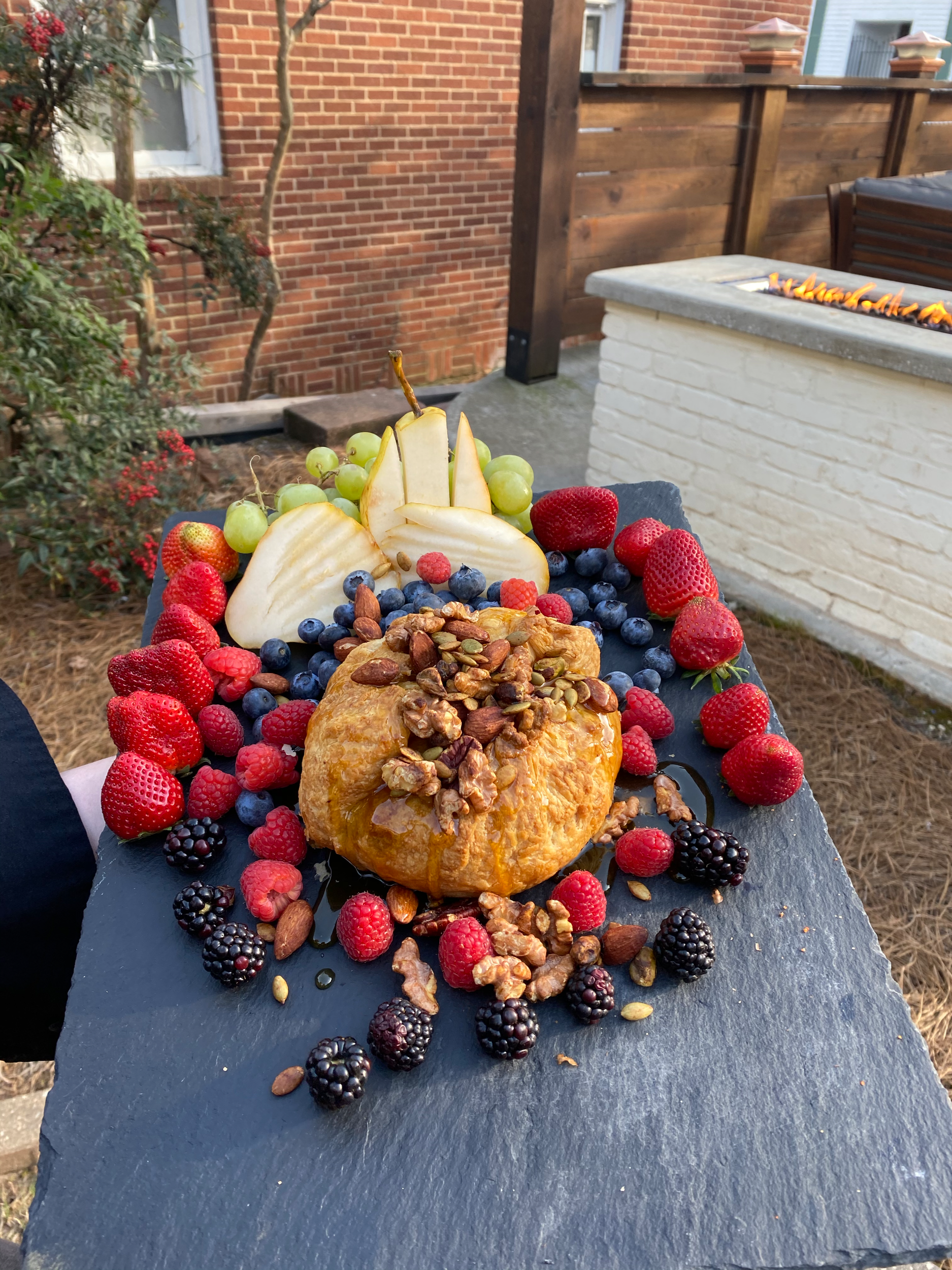 A dessert with mixed berries, sliced pears, green grapes, a pastry topped with nuts and seeds, honey, and a dark slate serving board.