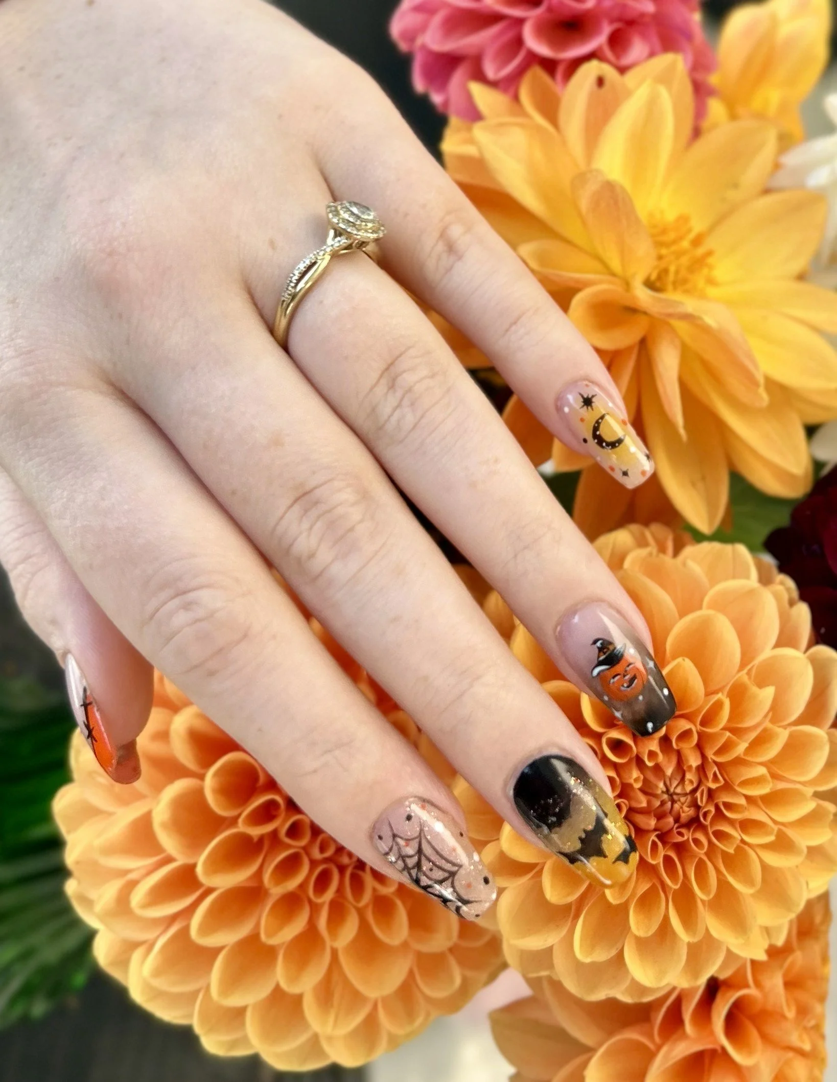 Hand with Halloween-themed nail art resting on orange and pink dahlias. The nails feature designs like a spider web, pumpkin, witch hat, moon, and stars. The hand is wearing a gold ring with a diamond setting.