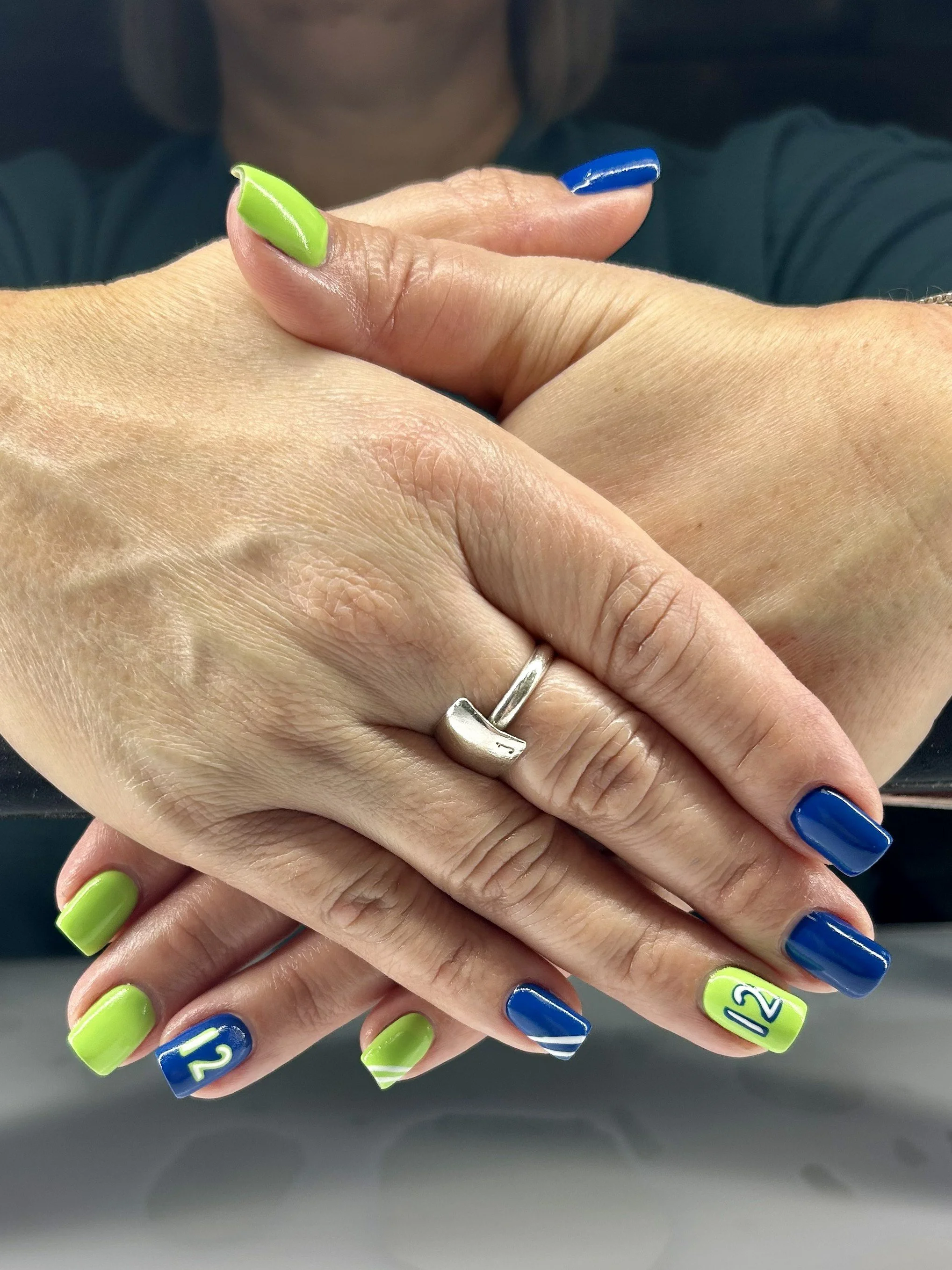 Person’s hands with colorful nail art, one hand with a ring, crossed over each other, reflected in a mirror