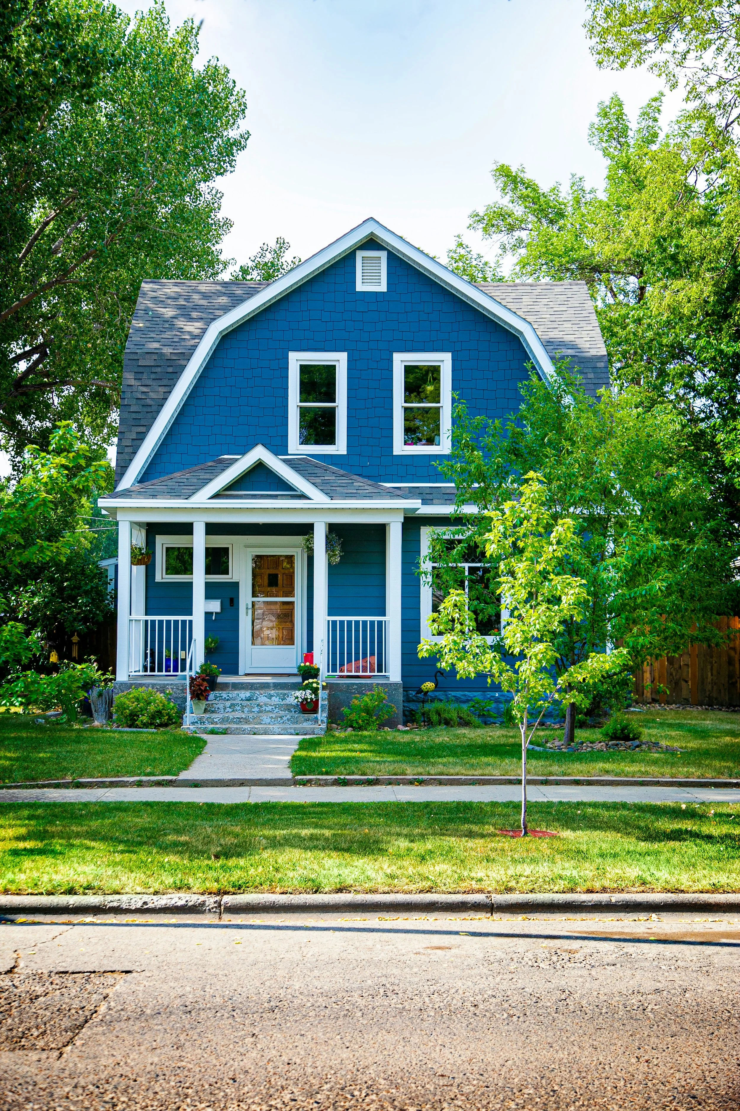 A large, two-story house with a wraparound porch, shingled exterior siding, black shutters, multiple windows, and a manicured lawn surrounded by trees.
