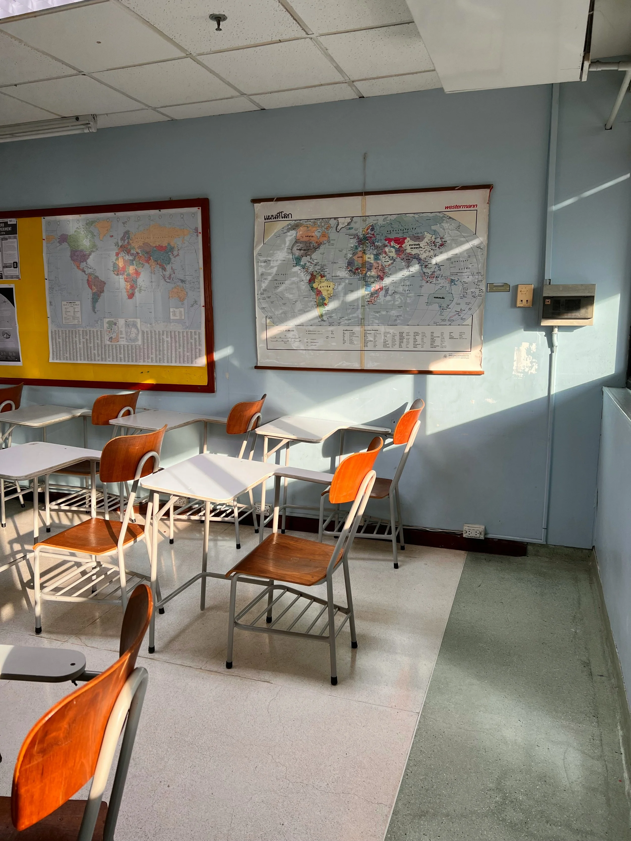 Empty classroom with wooden and metal desks and chairs, two world maps on light blue walls, sunlight casting shadows.