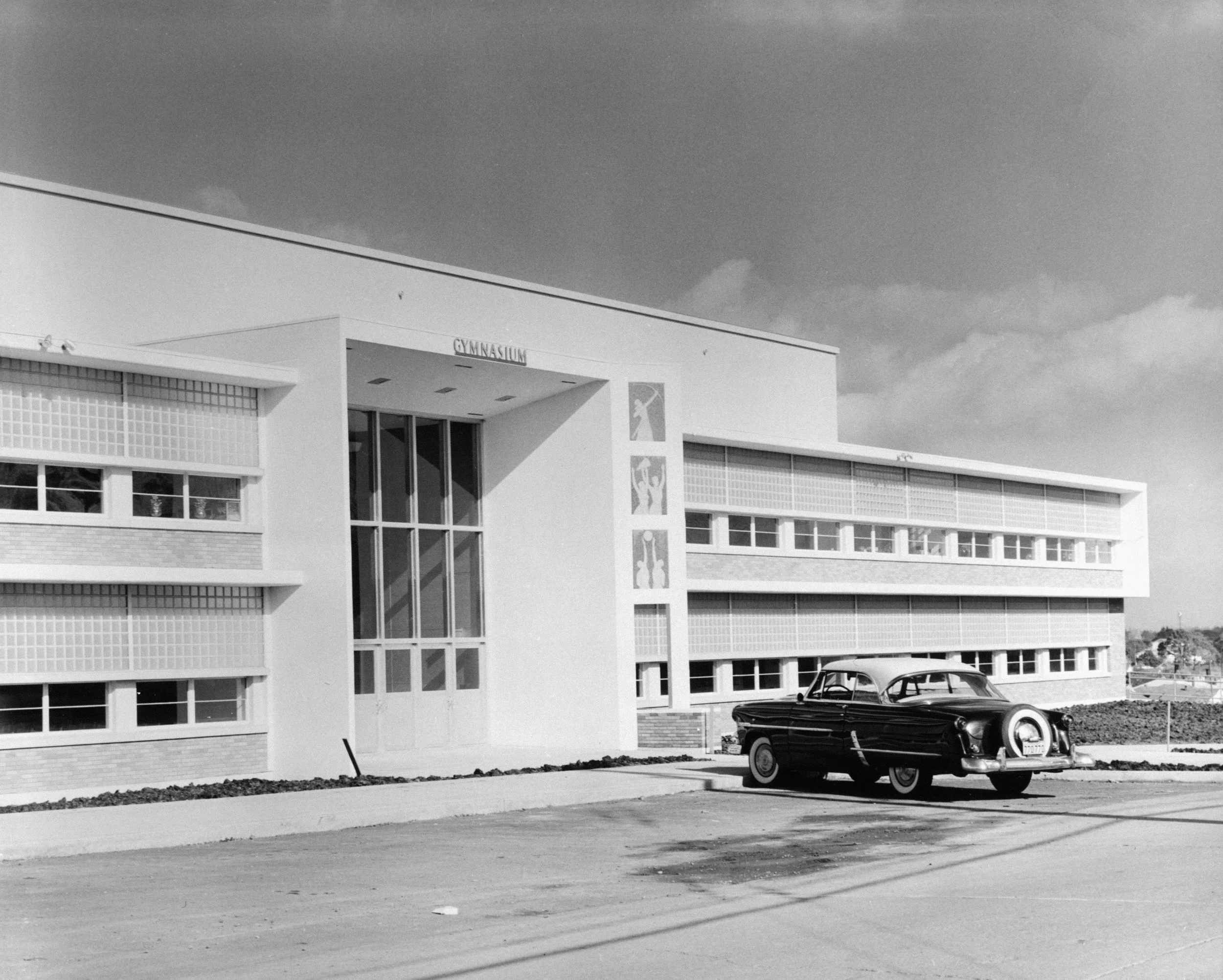 Black and white photo of a modernist school building with the sign 'Gymnasium' and a vintage black car parked in front.