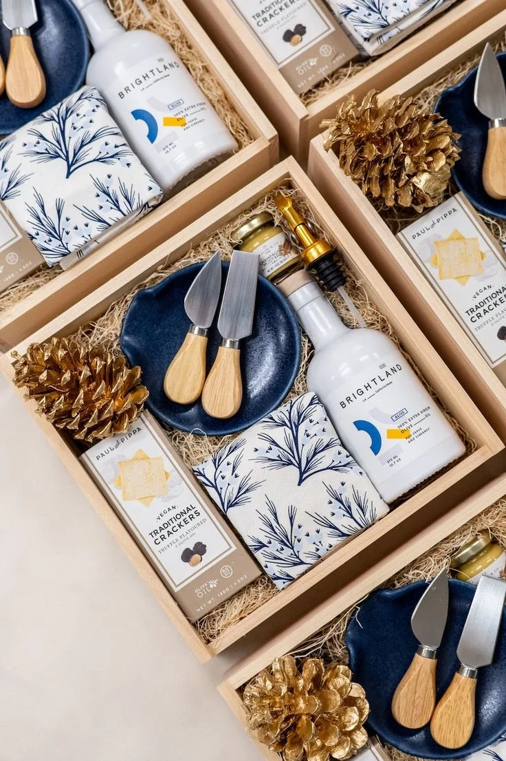 Gift boxes containing kitchen items and snacks, including a set of cheese knives on a blue plate, a white bottle labeled 'Brightland,' gold pinecones, black and white patterned napkins, cracker packages, and a gold honey dipper.