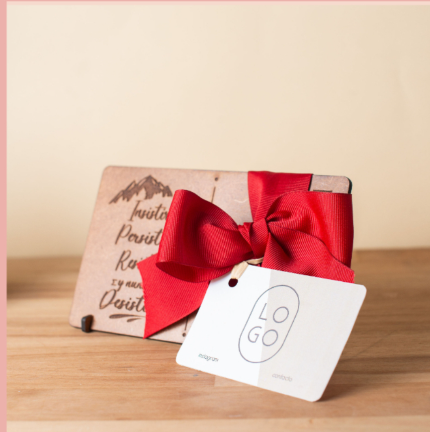 A gift card wrapped with a large red ribbon bow, placed on a wooden surface in front of a wooden plaque with inscriptions and mountain graphic.