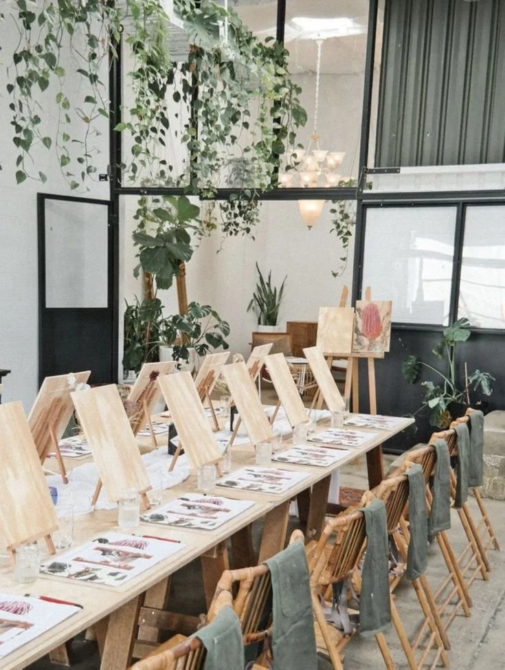 A long table set up for a painting class with individual easels, wine glasses, and menus, inside a bright room decorated with hanging plants and framed artwork.