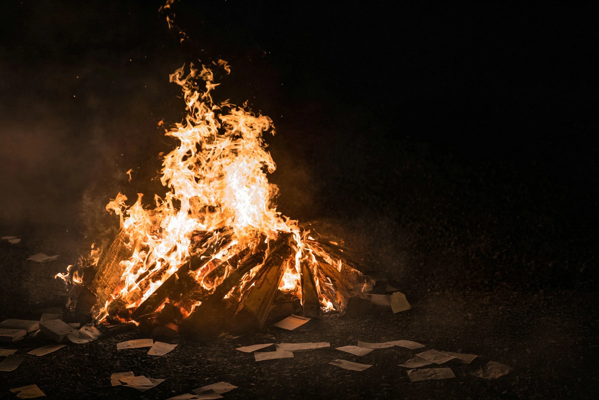 A large bonfire with burning wooden logs and scattered papers on the ground at night.