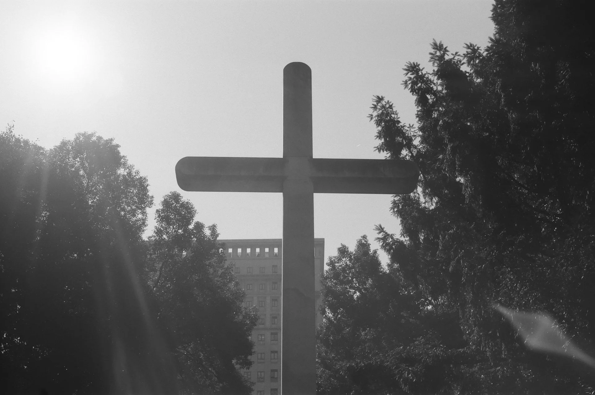 Black and white photo of a large cross-shaped monument surrounded by trees with sunlight shining through.