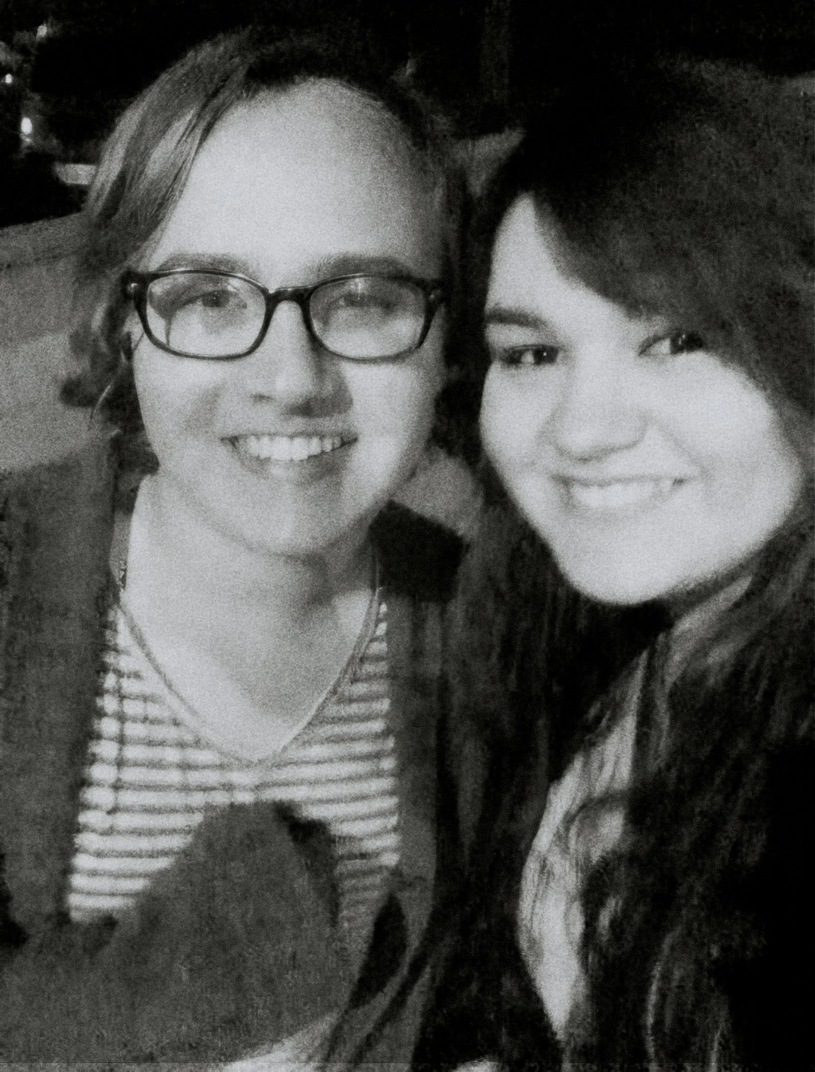 Two young people smiling closely together in black and white, one with glasses and light hair, the other with dark hair.