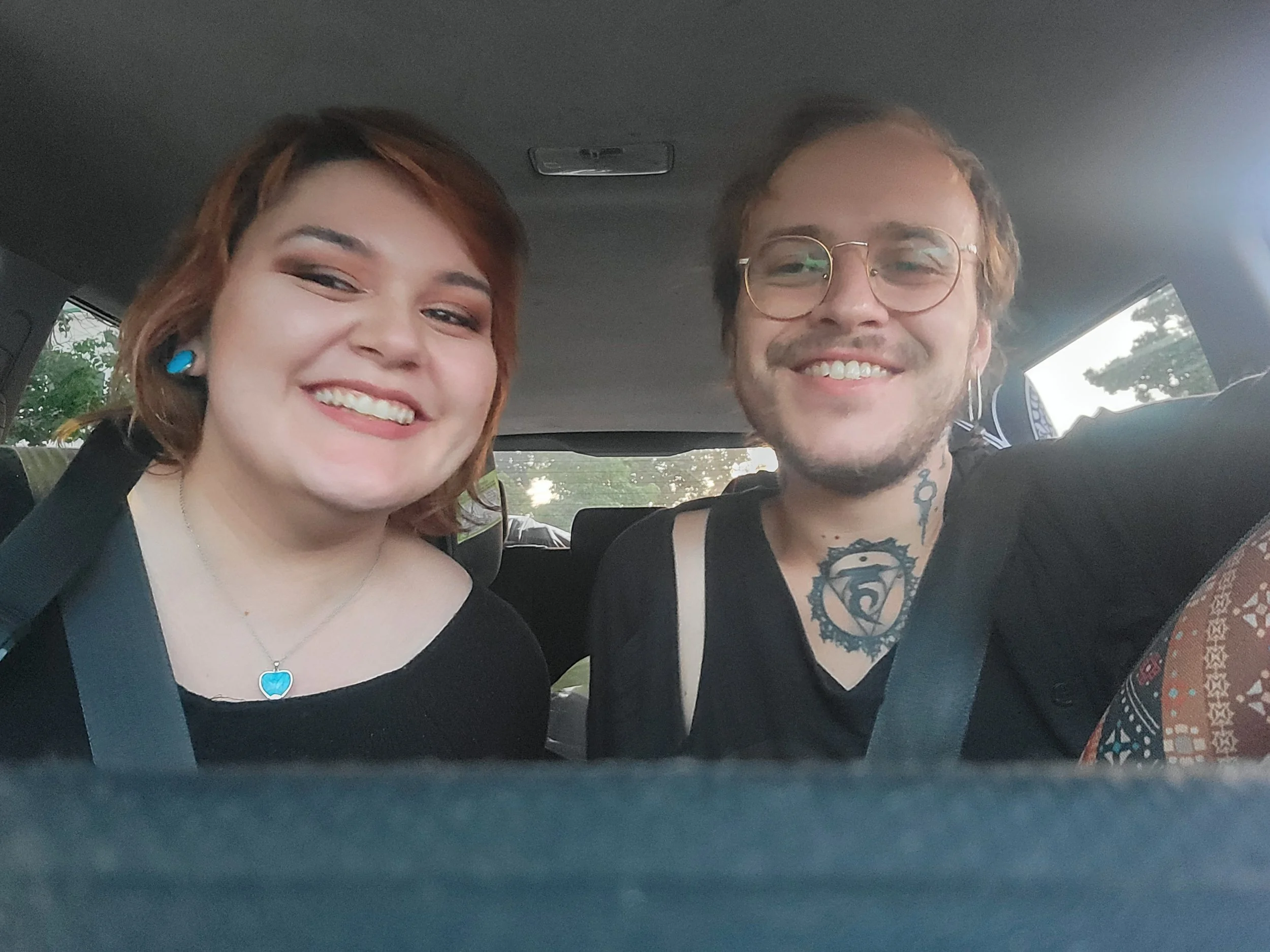 Smiling woman with short red hair and a man with glasses and tattoos inside a car.