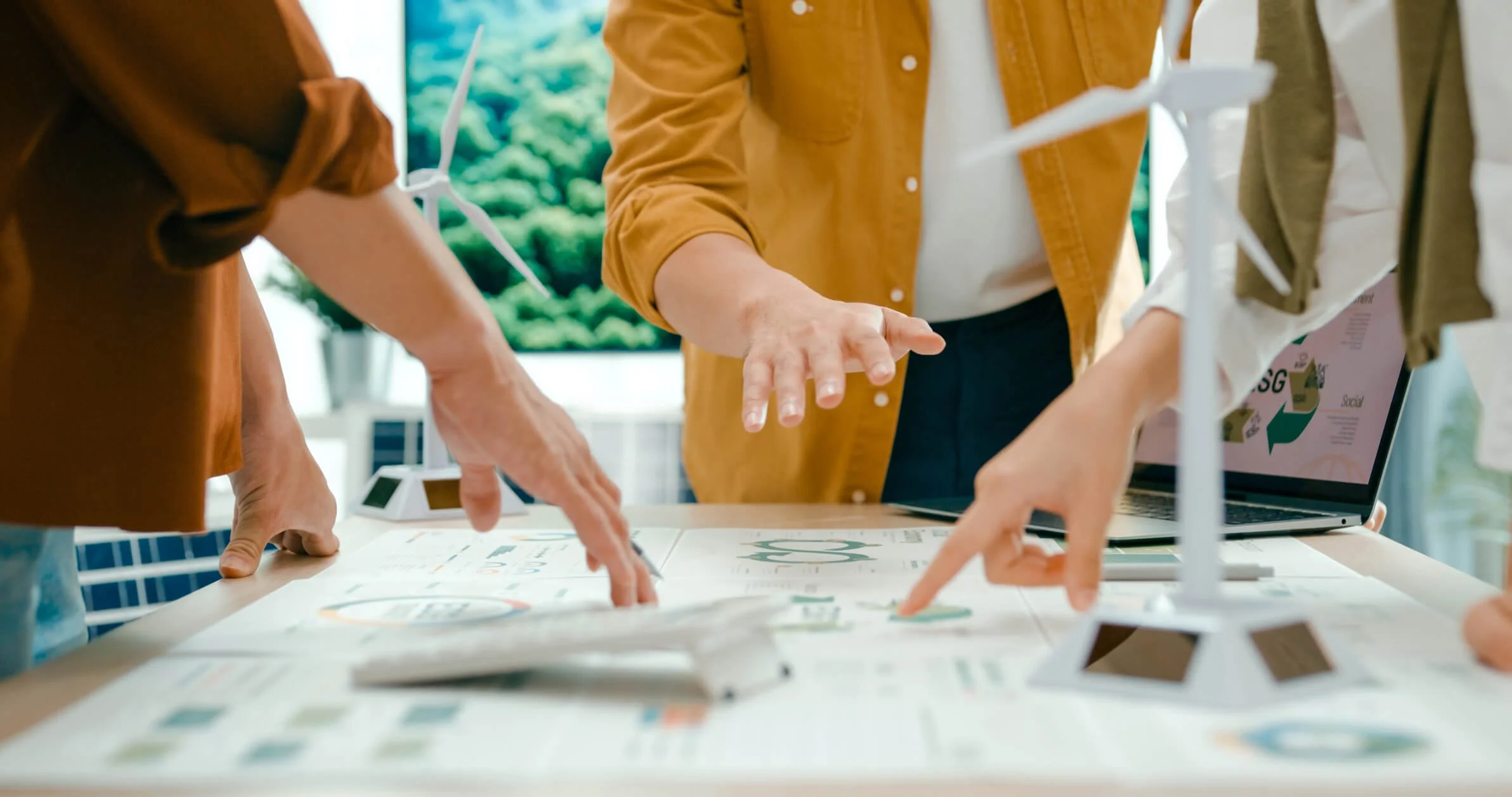 People working on a table with a laptop and wind turbine models, discussing environmental sustainability.