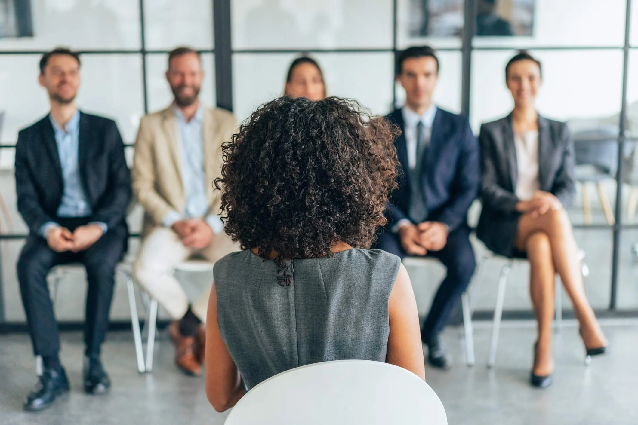 A woman with curly hair sitting in front of a panel of five professionals in suits, during an interview or presentation in a modern office setting.