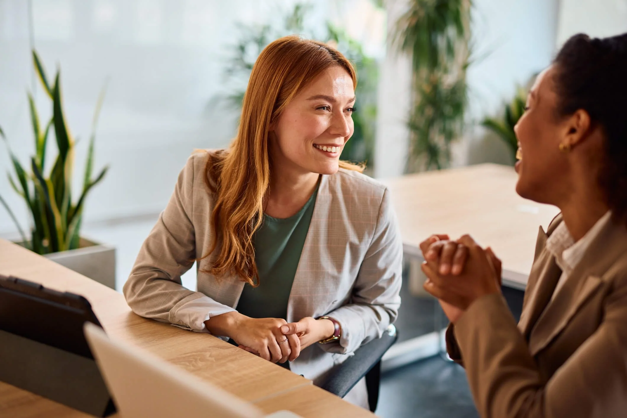 Two women sitting at a desk in a bright office, engaging in a conversation and smiling.