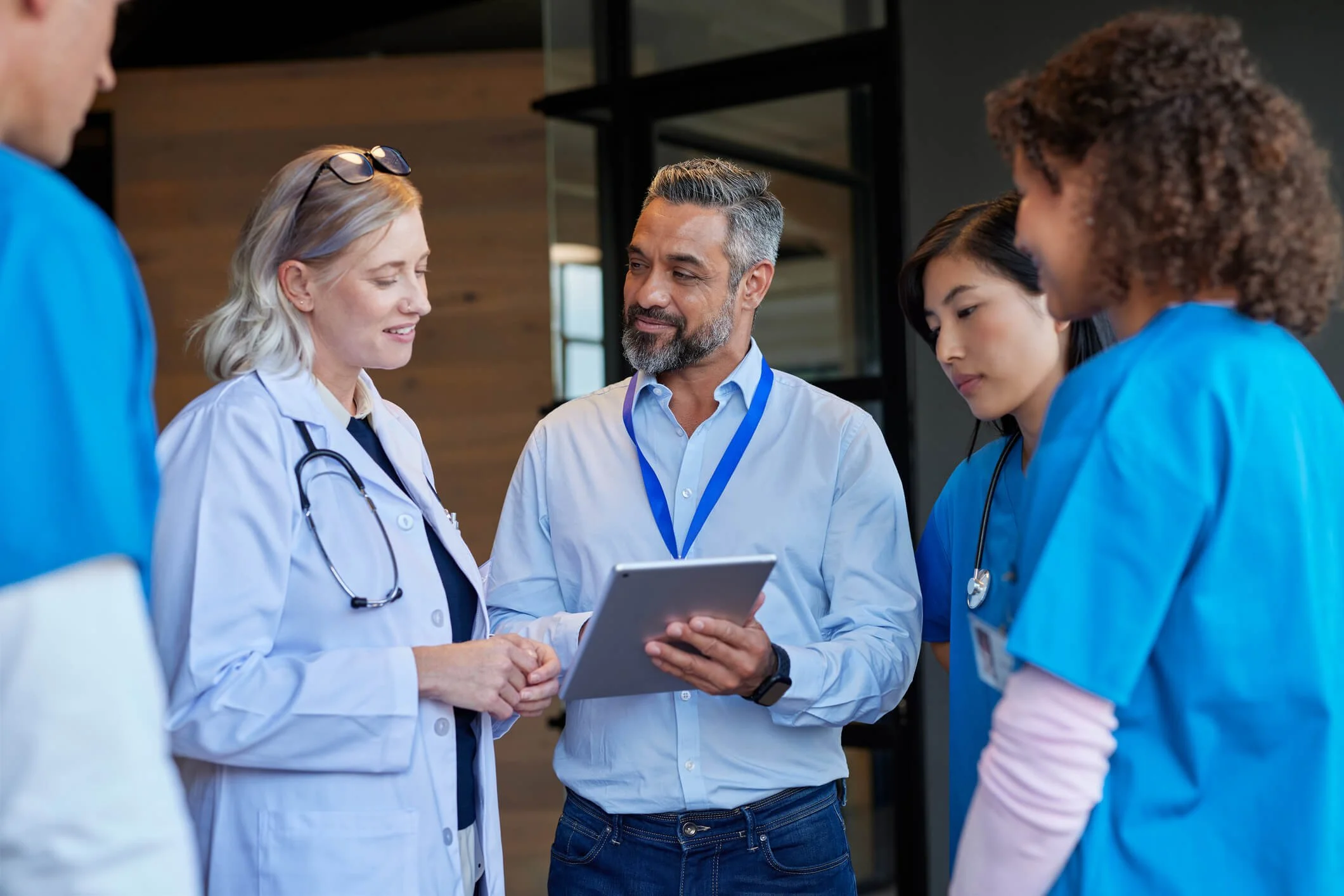 A group of healthcare professionals, including a doctor with a stethoscope and medical staff in scrubs, are engaged in a discussion with a man holding a tablet in a hospital or medical facility.
