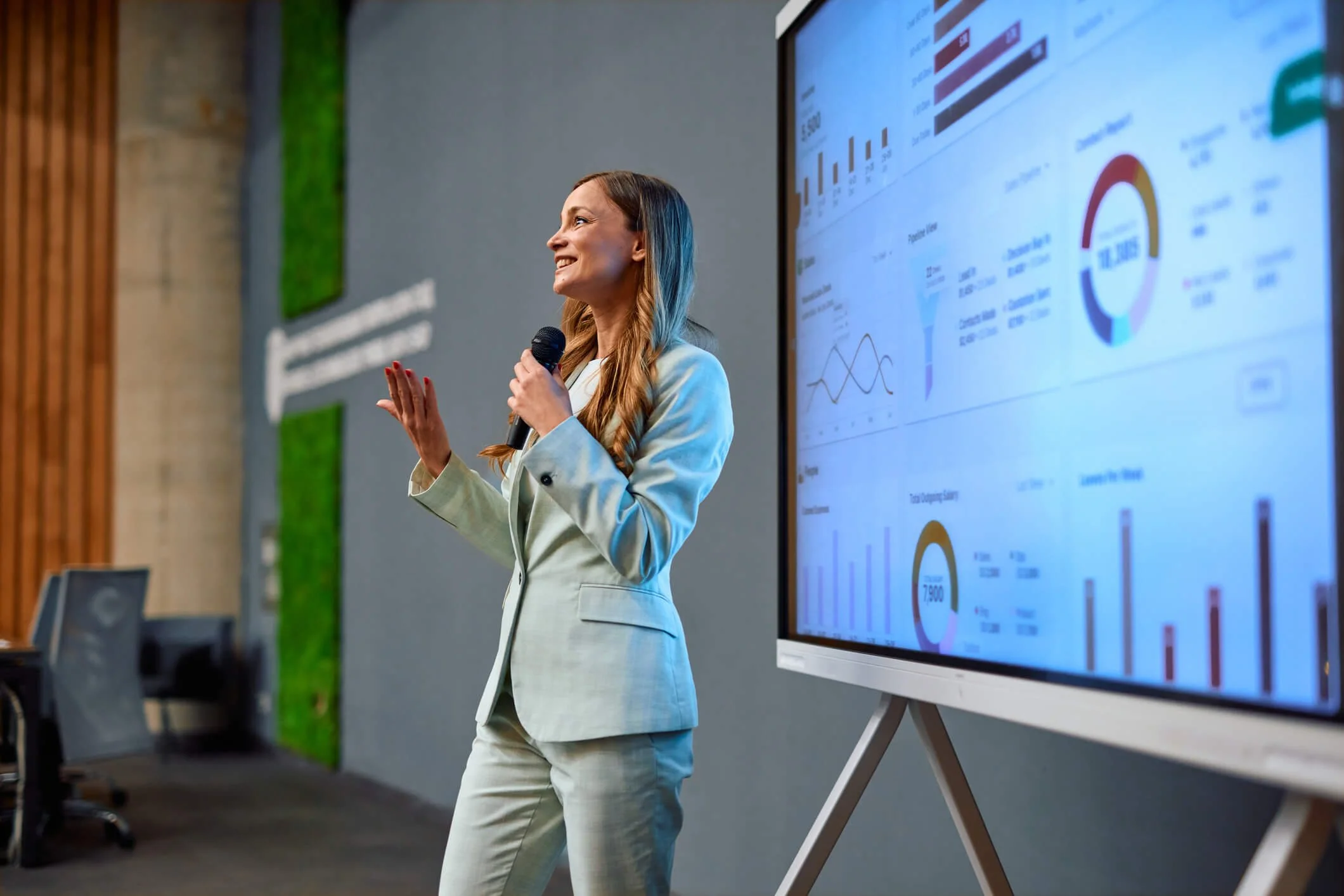 A woman in a light-colored business suit giving a presentation with bar graphs, pie charts, and line graphs on a large digital screen in a conference room.