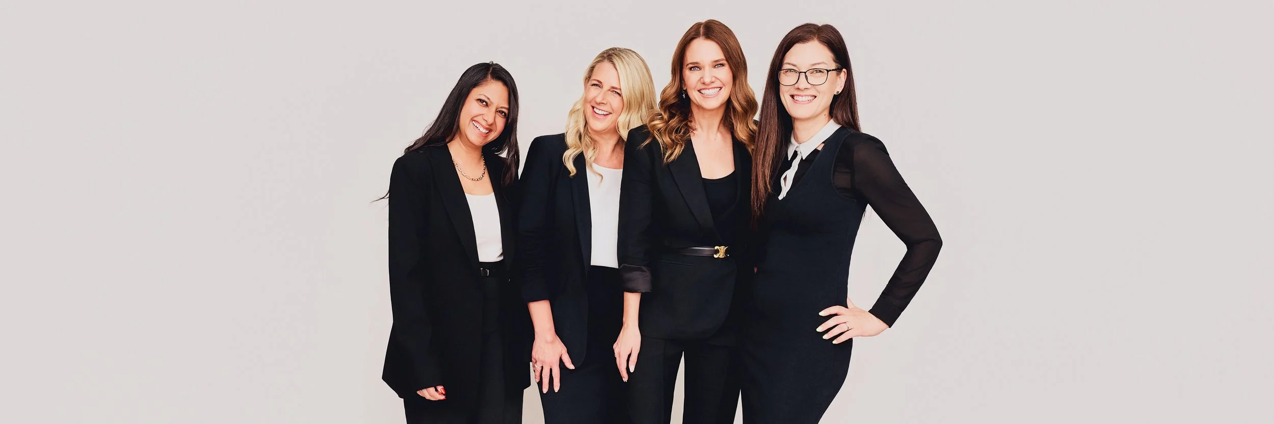 Four smiling women in professional black and white business attire standing against a plain light gray background.