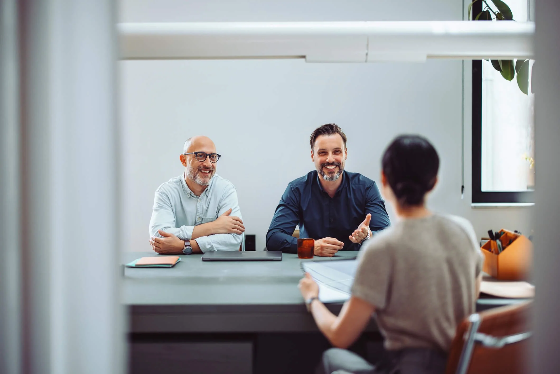 Two men and a woman sitting at a conference table during a meeting in a well-lit office, with a window and a potted plant in the background.