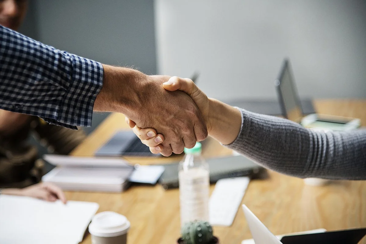 Two people shaking hands in a meeting room with laptops, notebooks, and a water bottle on the table.