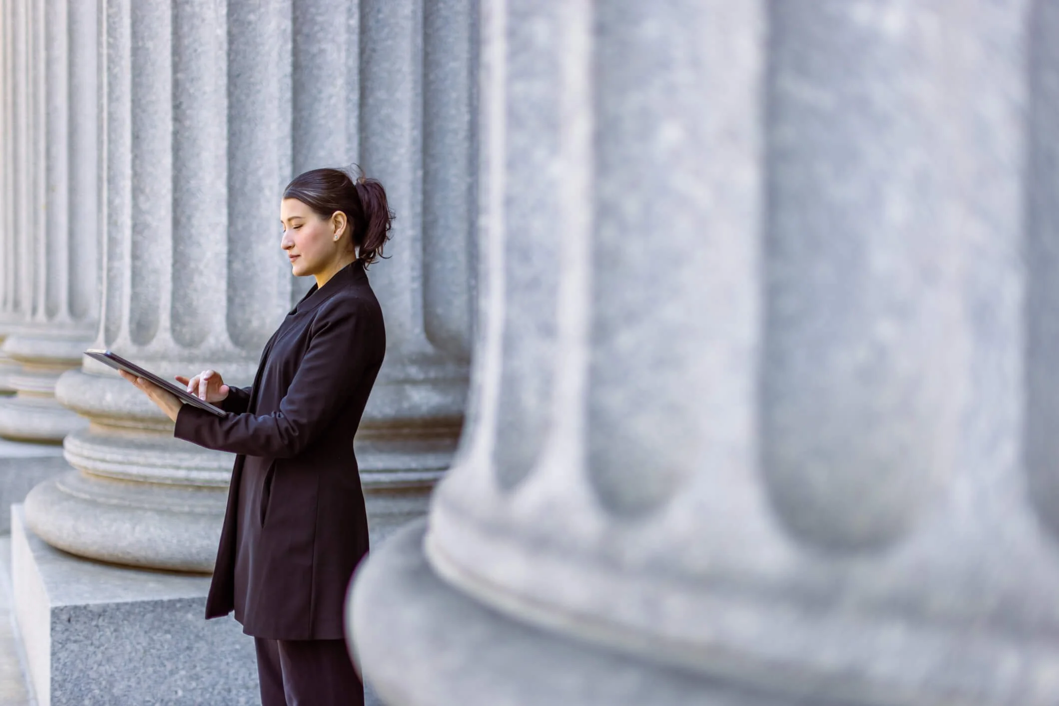 A woman in a black blazer using a tablet while standing next to large stone columns of a government or historic building.