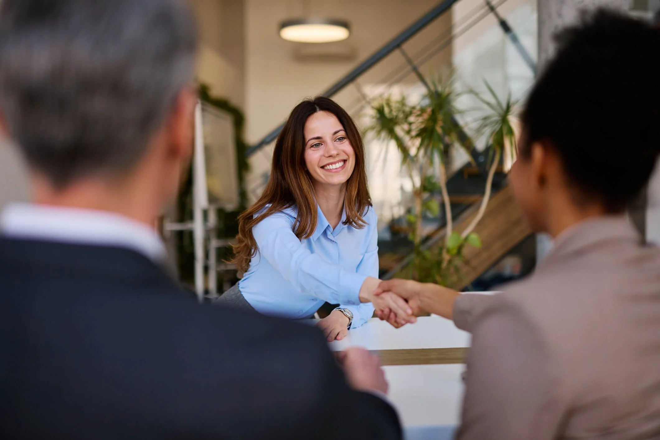 A woman with long brown hair wearing a light blue shirt smiling and shaking hands with a woman with black hair in a gray blazer in an office or modern indoor setting.