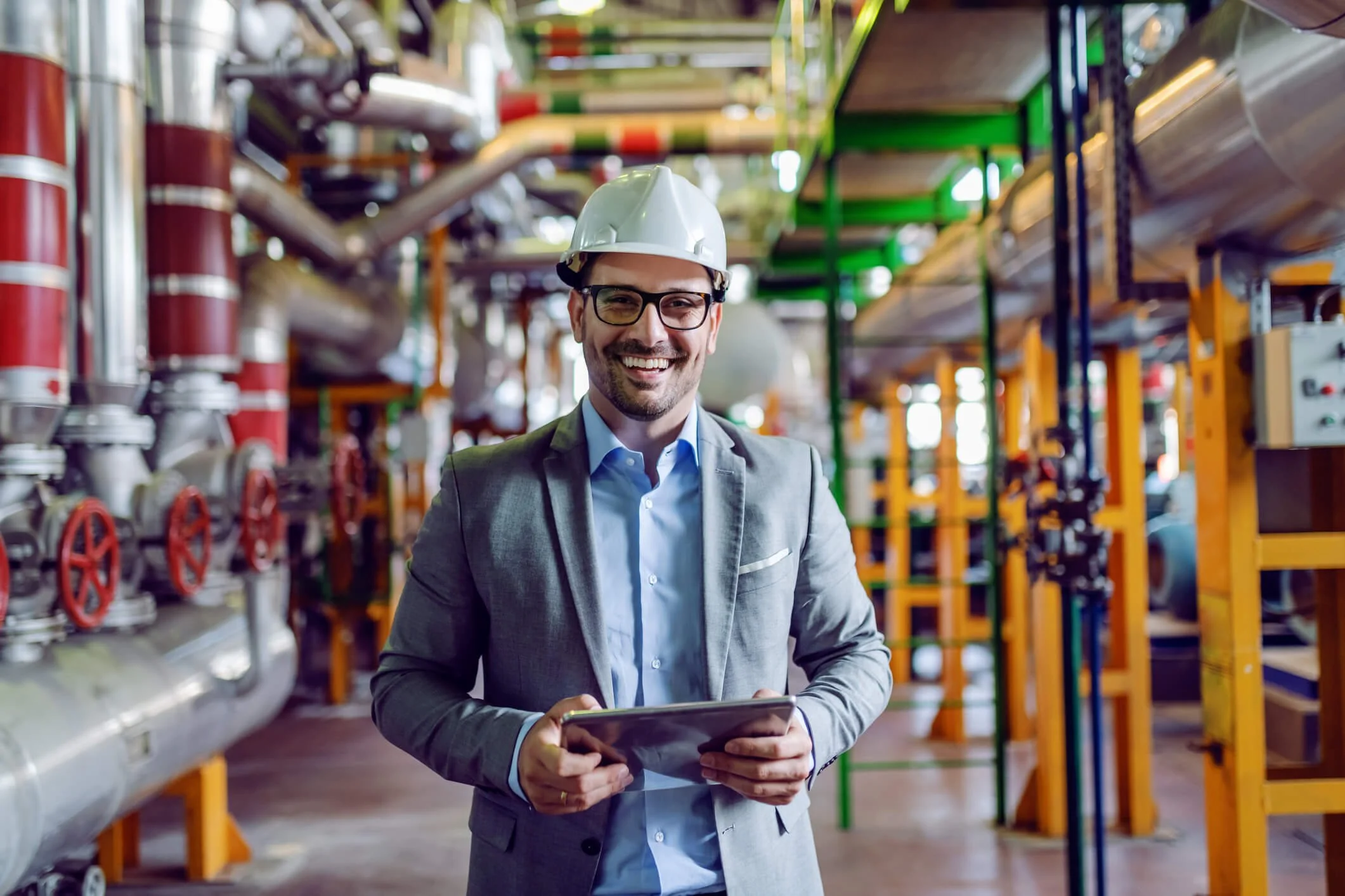A smiling man in a suit and hard hat holding a tablet in an industrial facility filled with pipes and machinery.