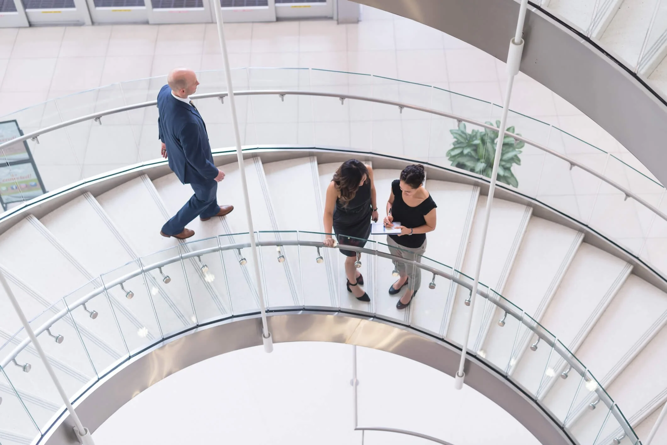 Two women and one man standing on a spiral staircase inside a modern building. The women are engaged in a discussion, with one showing notes on a clipboard. The man is walking down the stairs.