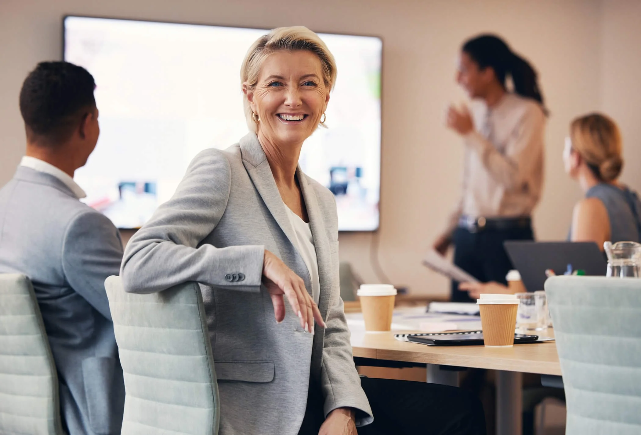 A smiling blonde woman in a gray blazer sits at a conference table with coffee cups, laptop, and notes, in a business meeting room with other professionals, two women and one man, in the background.