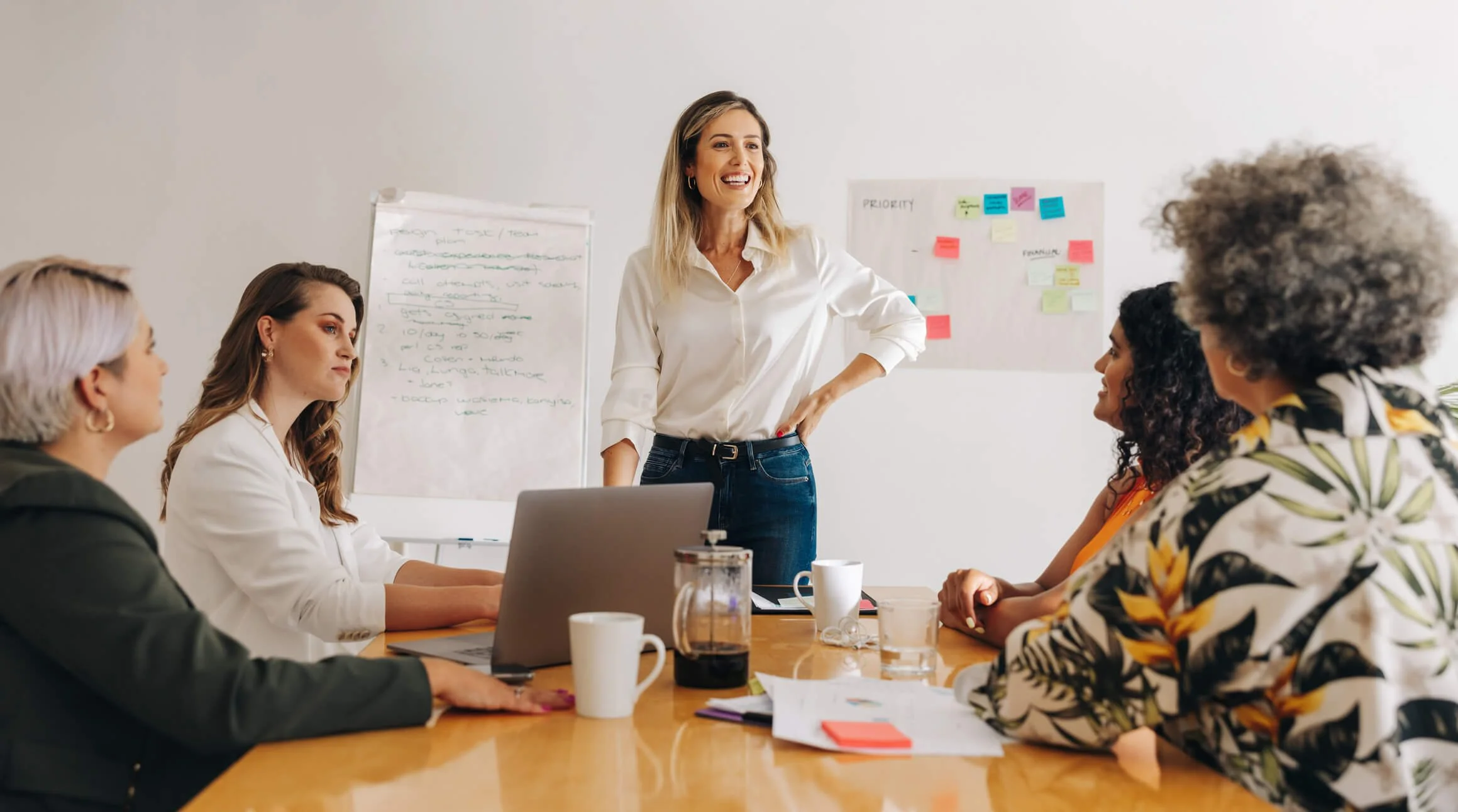 A woman standing and smiling during a meeting with four seated women at a conference table with laptops, coffee cups, and papers, in a bright office room with white walls and a whiteboard with notes.