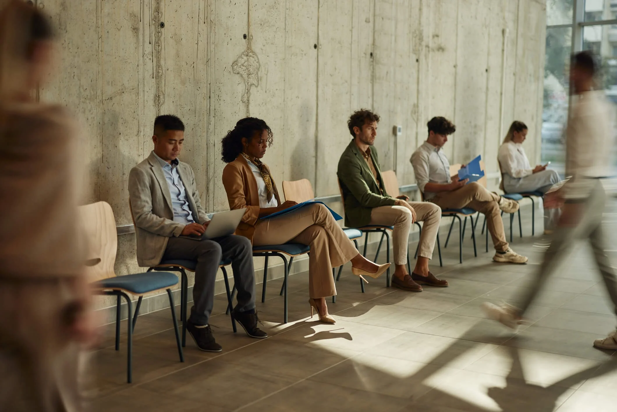 People sitting in a waiting area with a concrete wall, some using laptops or reading documents, and a person walking past in the foreground.