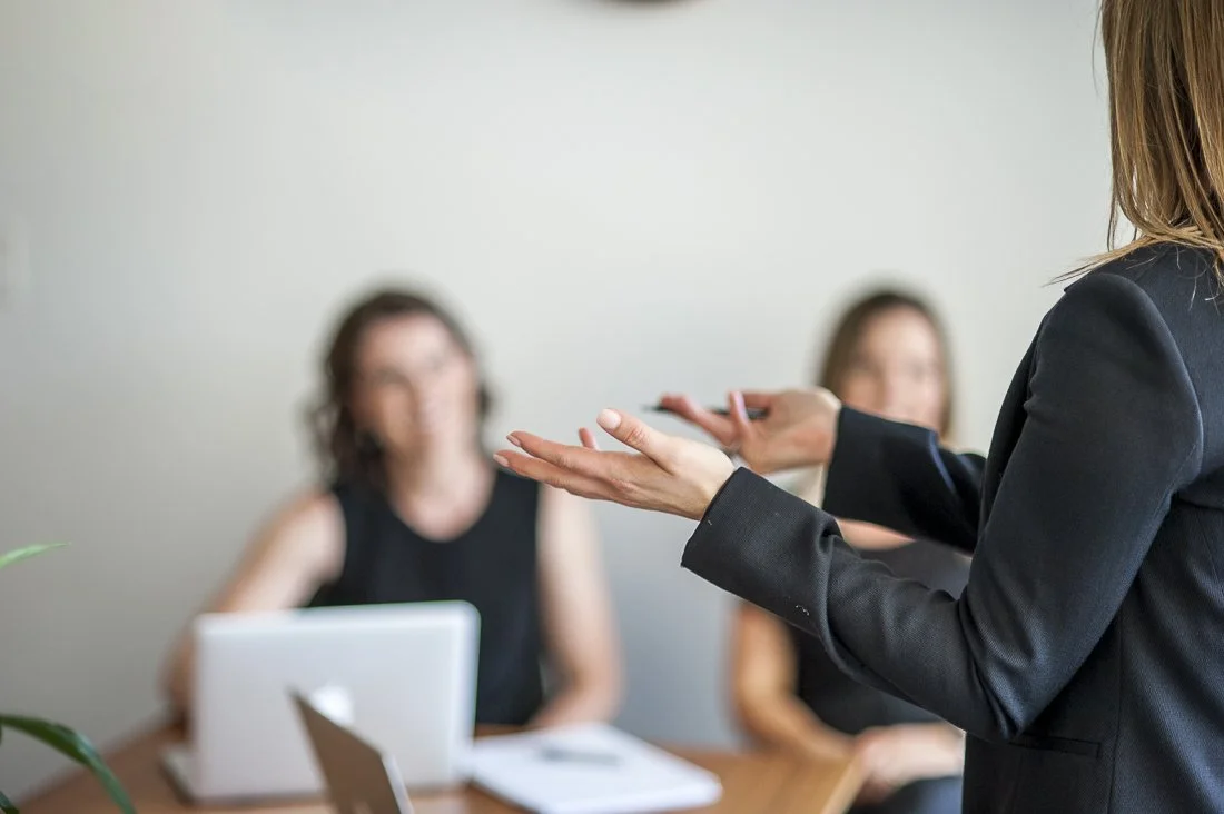 A woman in business attire giving a presentation to two seated women in a meeting room.