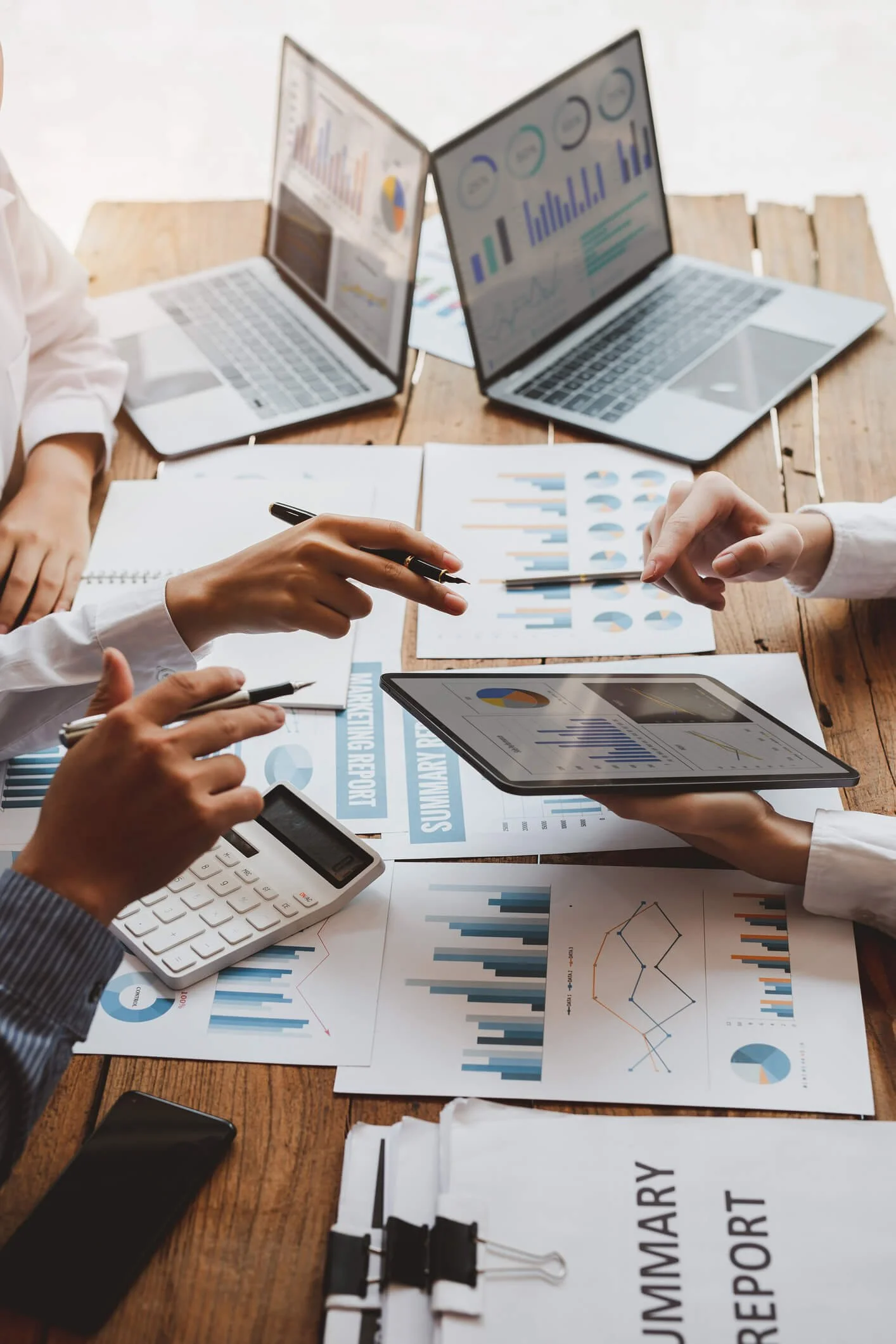 Business meeting with people analyzing financial charts and documents, using laptops, tablets, and a calculator on a wooden table.