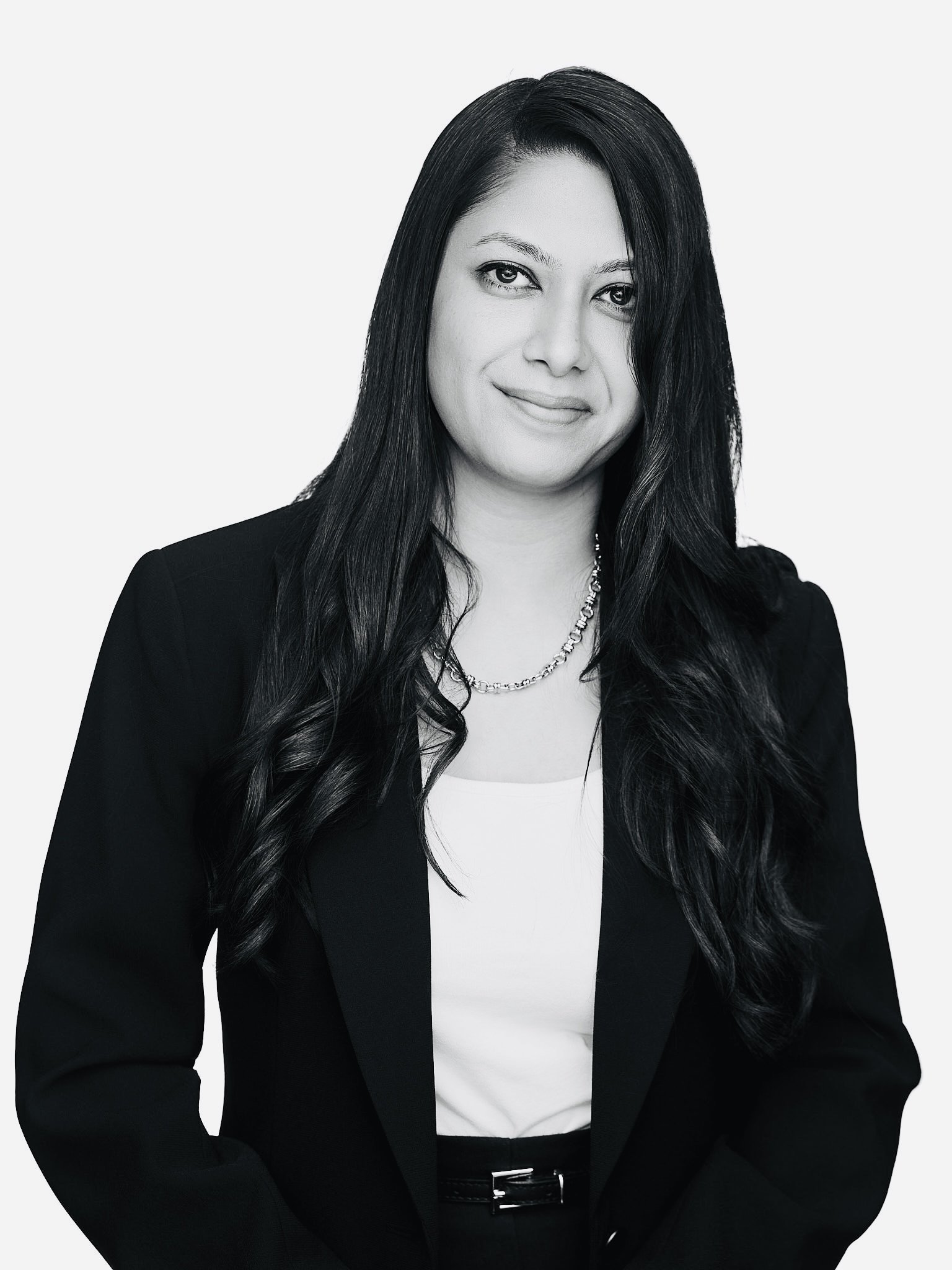 Black and white portrait of a woman with long, wavy dark hair, wearing a blazer, white top, and a necklace, smiling softly against a plain background.