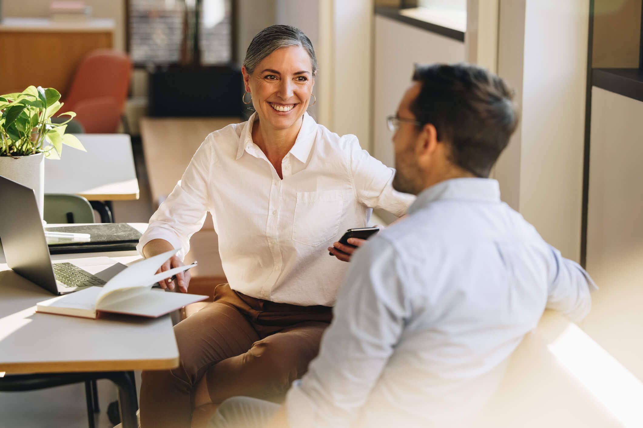 A woman and a man having a conversation in an office setting. The woman is smiling and holding a smartphone, while the man is sitting and looking at her. There is a laptop, open notebooks, and a plant on the table.