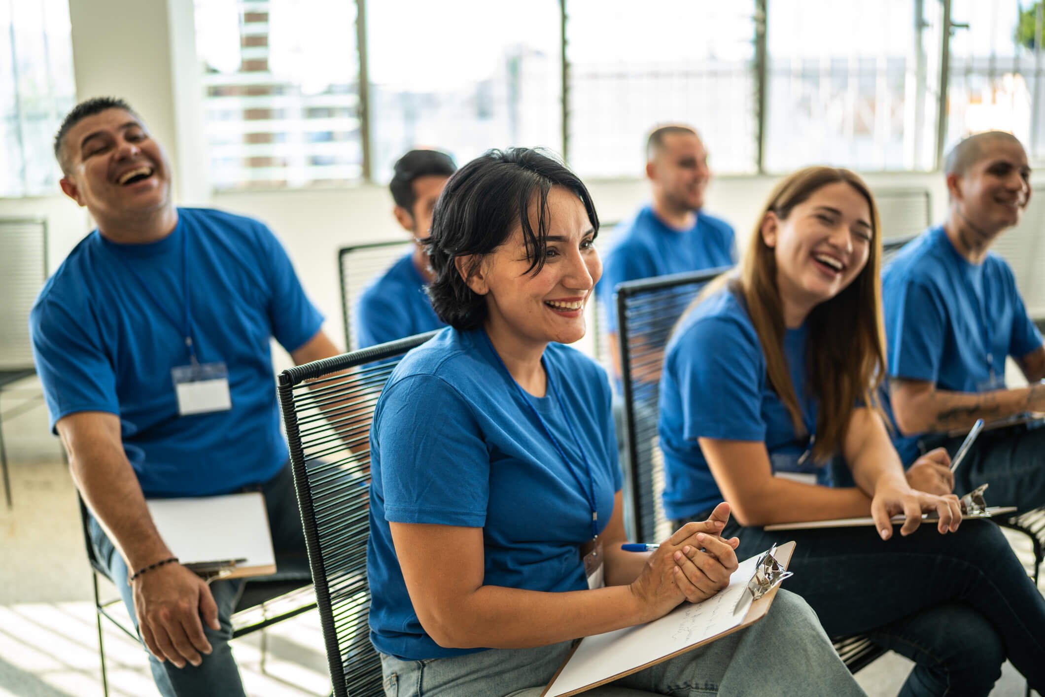 A group of diverse people wearing blue shirts, sitting in a classroom or conference room, smiling and laughing during a seminar or training session.