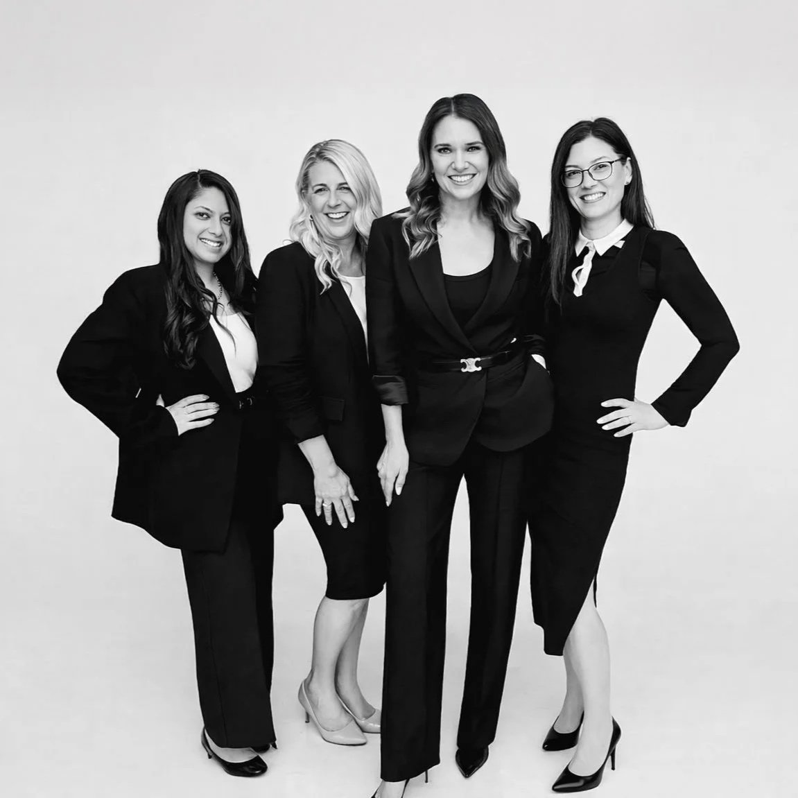Four women dressed in formal business attire standing together and smiling for a group photo against a plain background.
