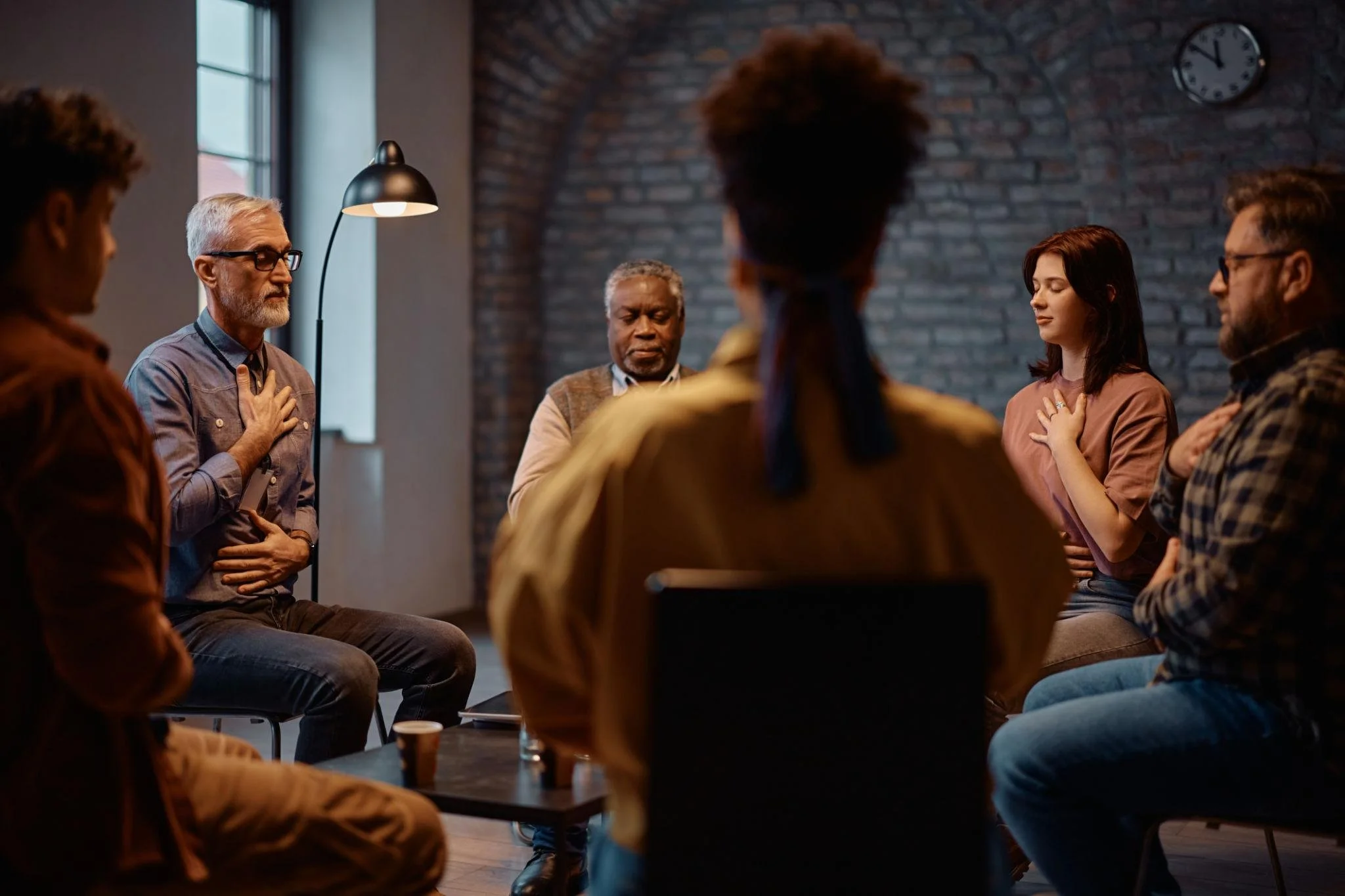 A group of diverse people participating in a meditation or prayer circle with their hands over their hearts, seated in a room with a brick wall and a clock on the wall.