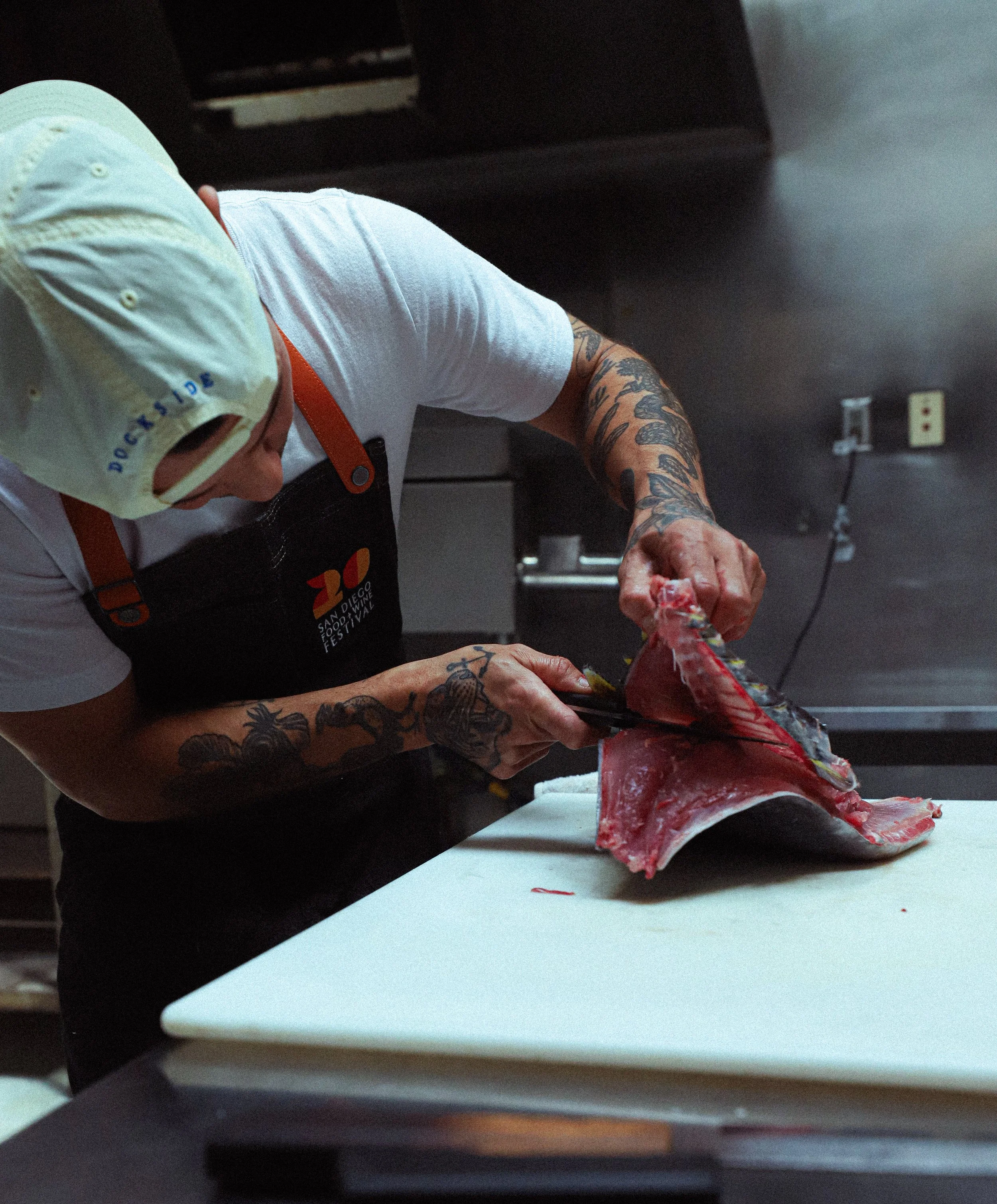 Person wearing a yellow cap and a black apron with 'San Diego Food Festival' logo, preparing to cut a large fish fillet on a white cutting board in a commercial kitchen.
