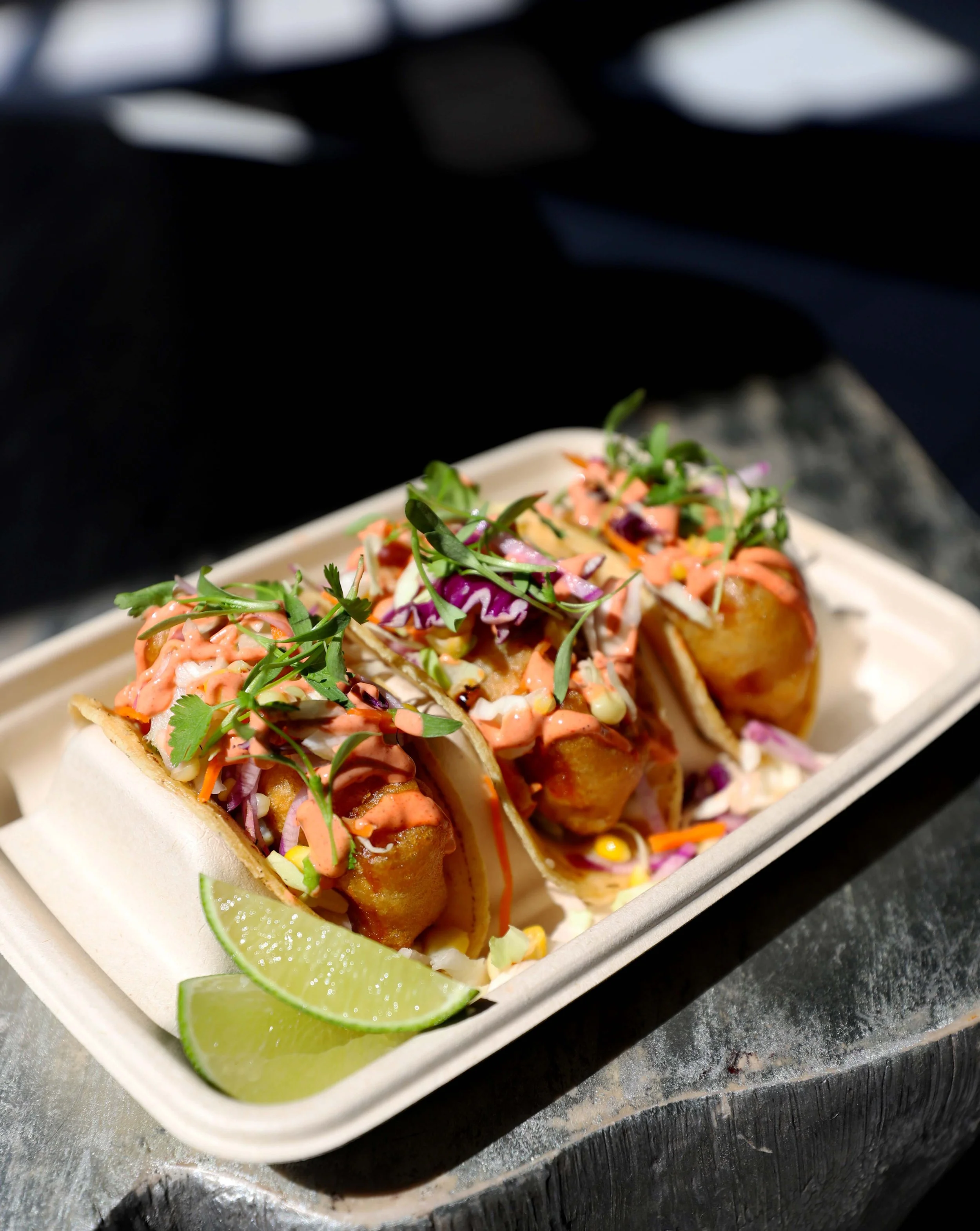 Three fish tacos topped with shredded cabbage, microgreens, and pink sauce, served with lime wedges on a white rectangular plate.