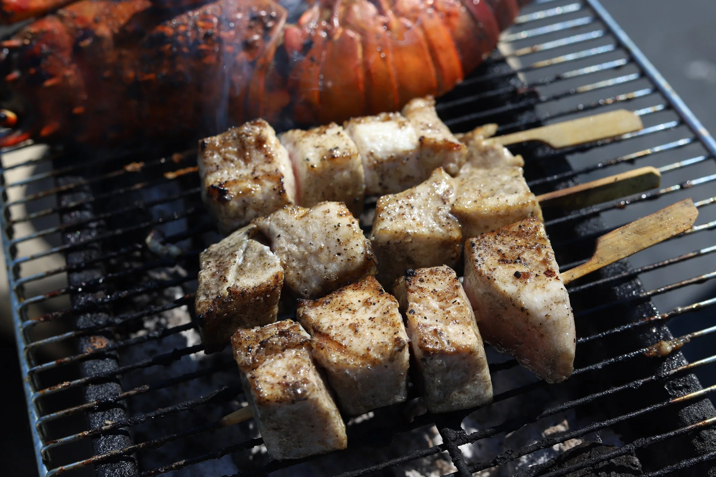 Cubes of seasoned swordfish belly cooking on a grill with a lobster in the background.