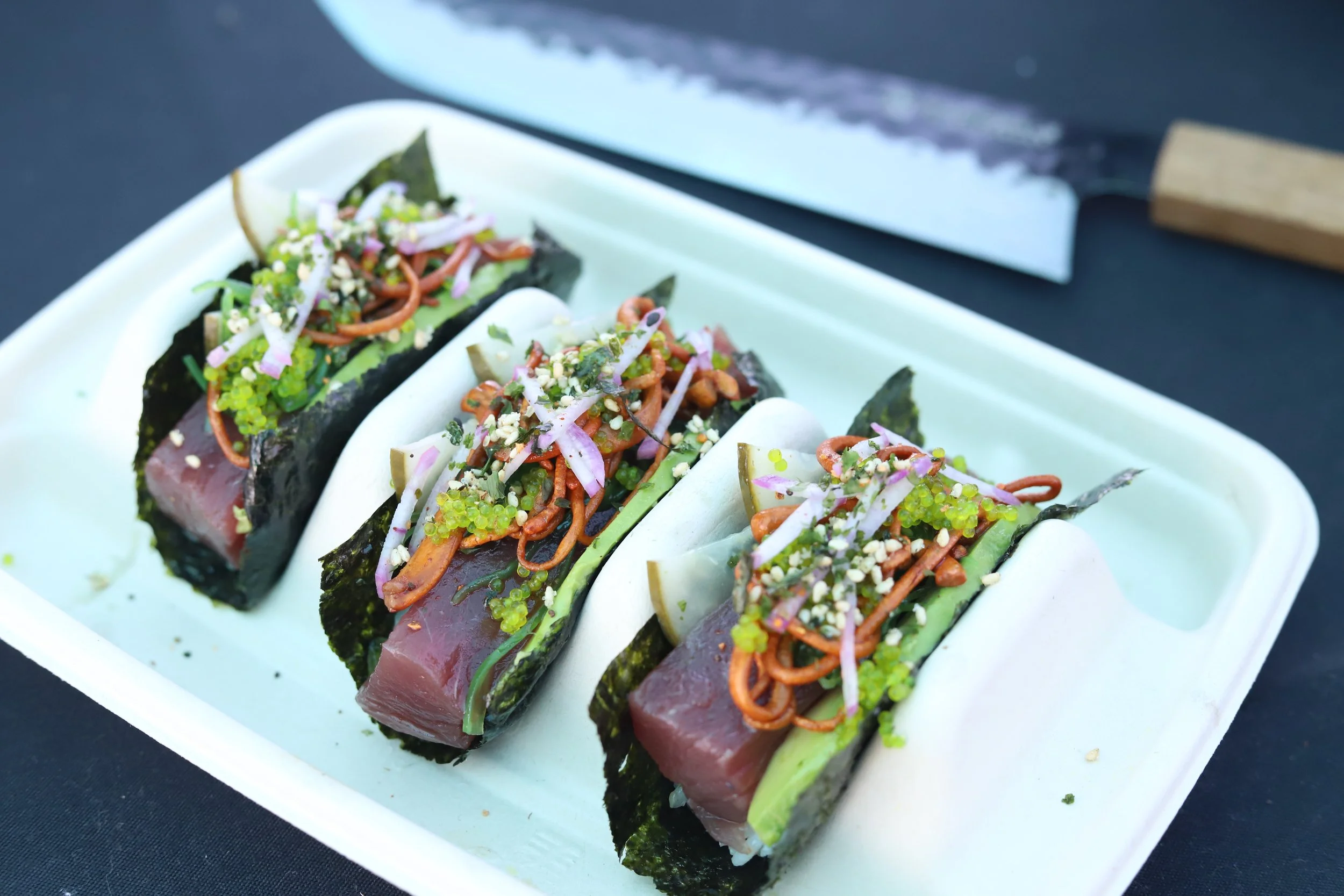 Three sushi tacos filled with raw fish, topped with chopped vegetables and sesame seeds on a white tray, with a knife in the background.
