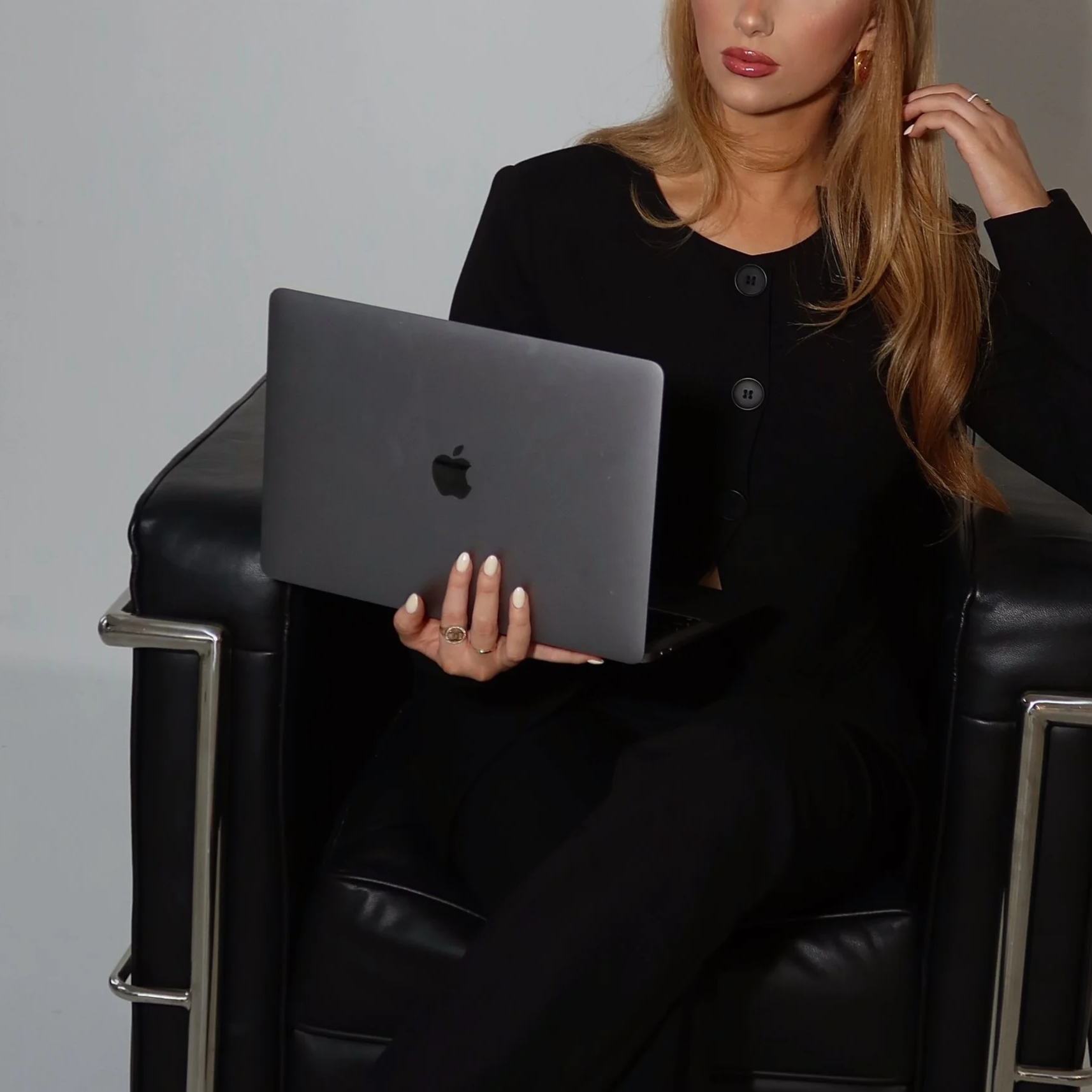 Woman with long blonde hair, red lipstick, gold earrings, and a black top sitting in a chair, holding a silver MacBook laptop.