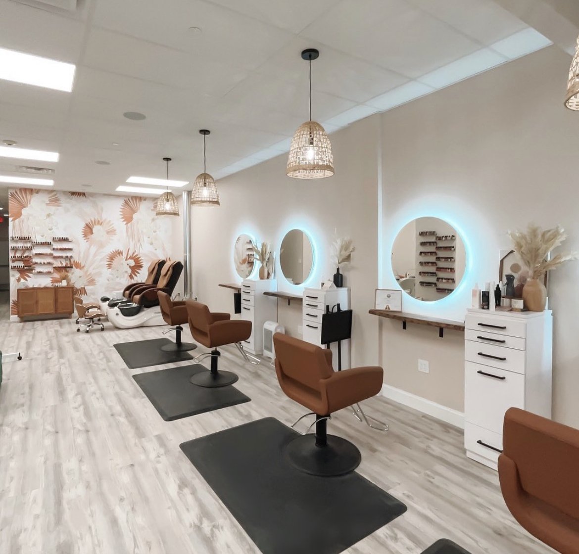 Interior view of a modern salon with three styling chairs, hair washing station, circular mirrors with backlit lights, and decorative vases with dried flowers.