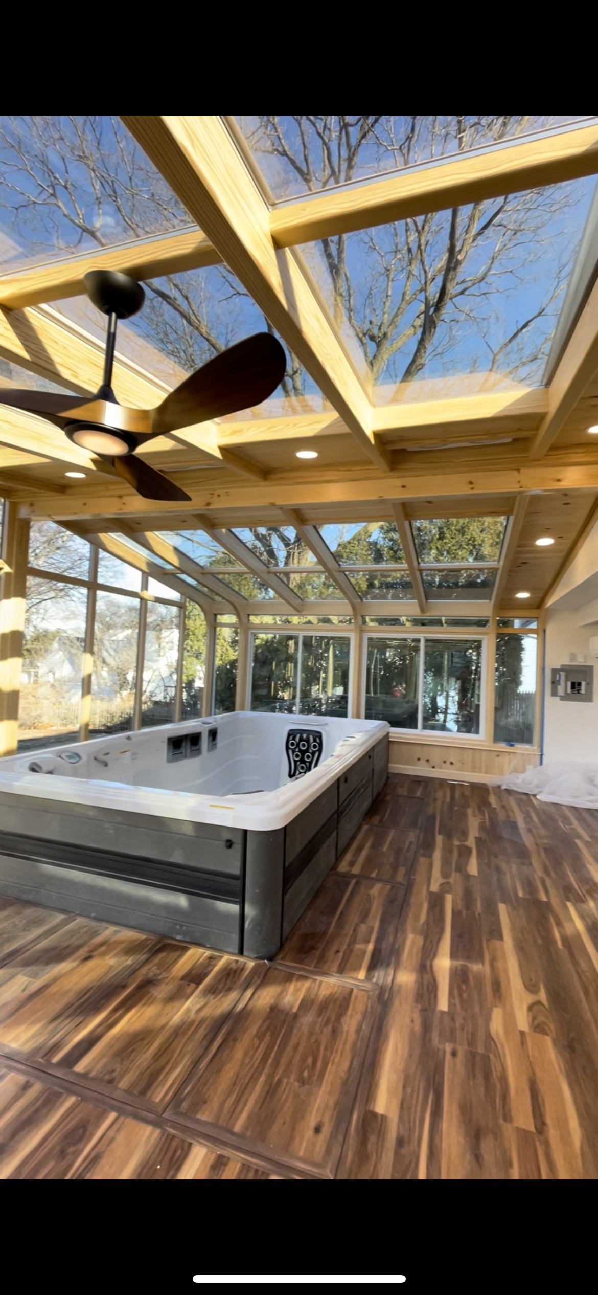 A sunlit room with a jacuzzi tub, wooden floors, large glass windows, a ceiling fan, and a glass ceiling showing trees and a blue sky outside.
