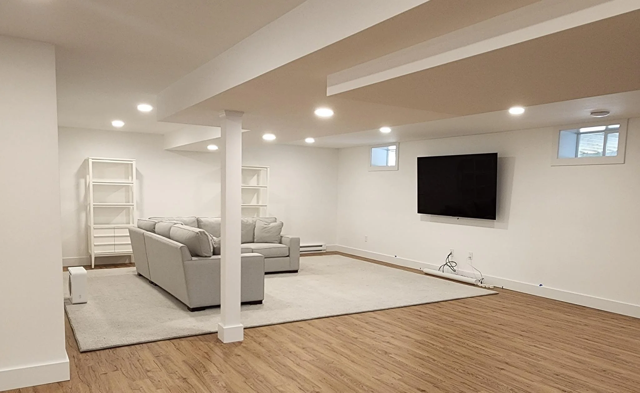 Empty living room with white walls, hardwood floors, a mounted flat-screen TV, a light-colored sofa, white bookshelves, a beige area rug, and small windows near the ceiling.